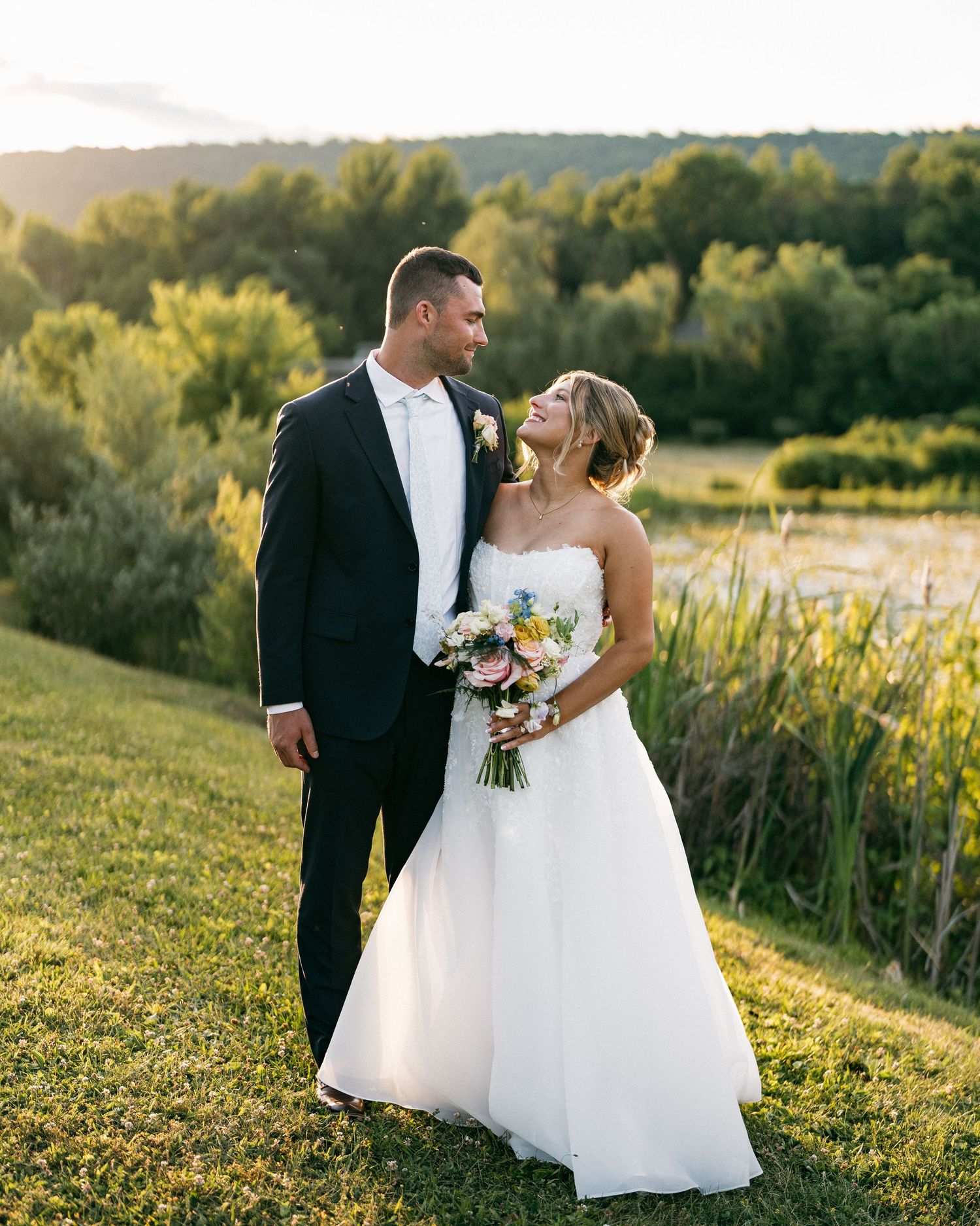 Bride and groom share a romantic moment at golden hour, standing on green grass with a scenic pond and trees behind them.