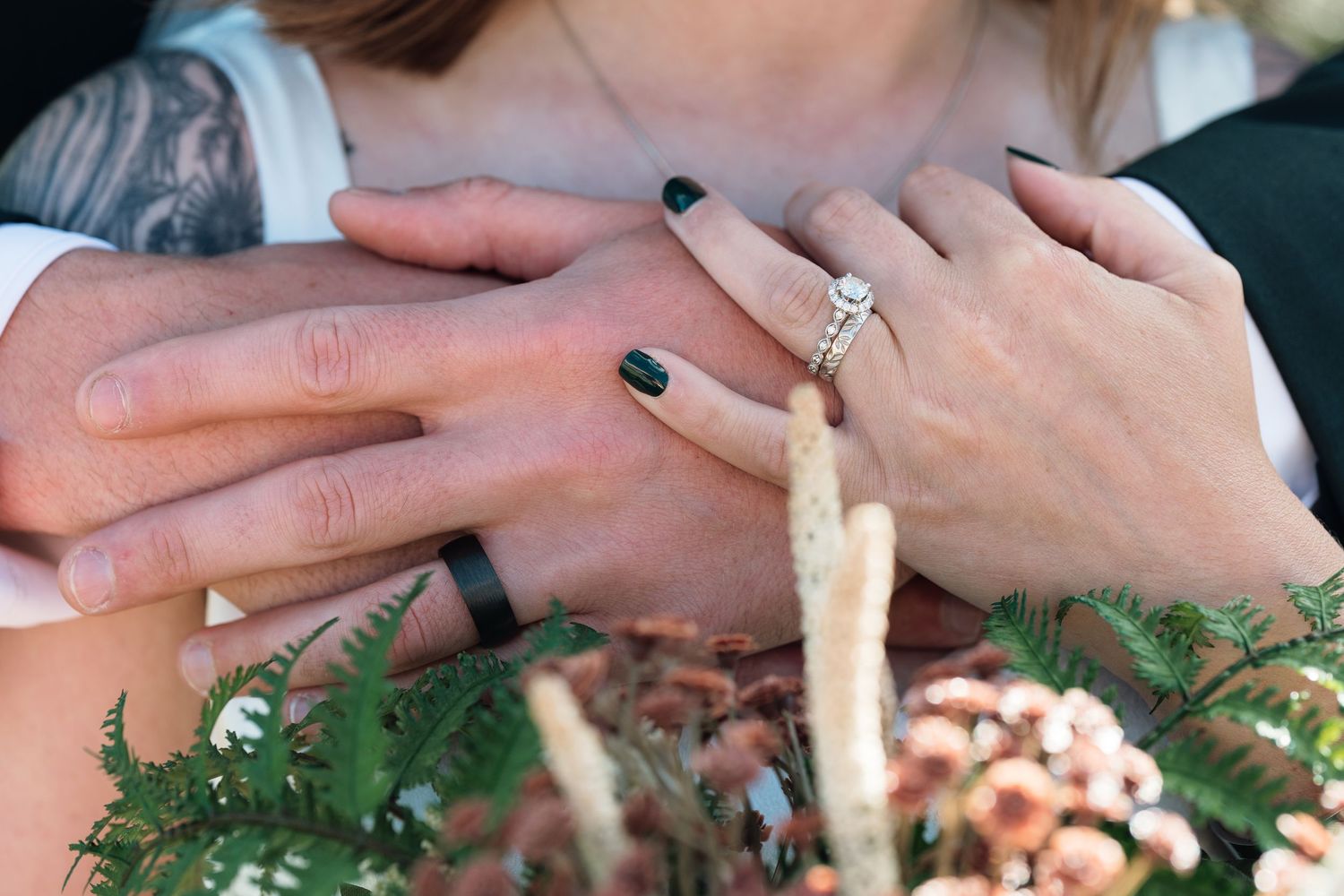 Newlyweds displaying wedding rings, woman wearing diamond engagement ring with black nails, man with black band, over floral bouquet.