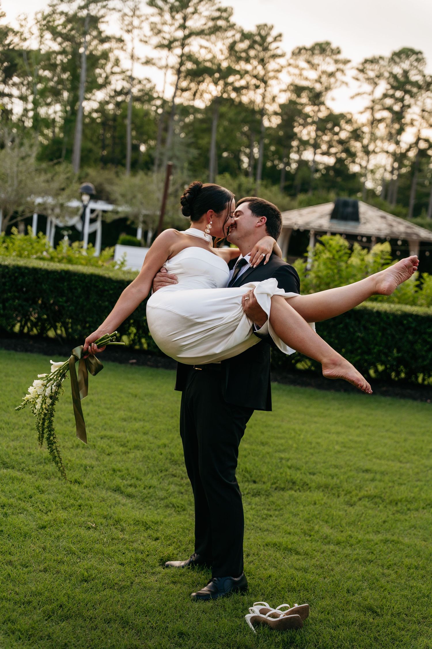 A romantic couple shares a kiss while performing a dramatic dip pose on a grassy lawn at sunset.