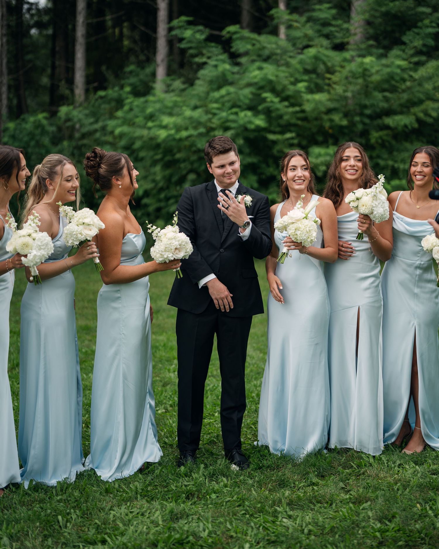 Groom in black suit surrounded by bridesmaids in light blue satin gowns holding white bouquets outdoors.