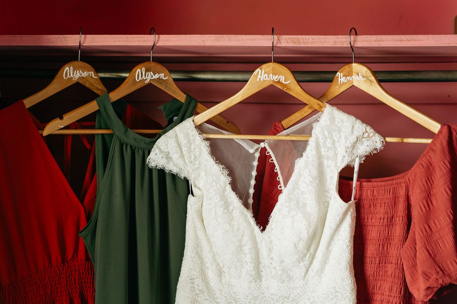 Wedding dresses and bridesmaid gowns in red and green colors hanging on wooden hangers against a red wall.