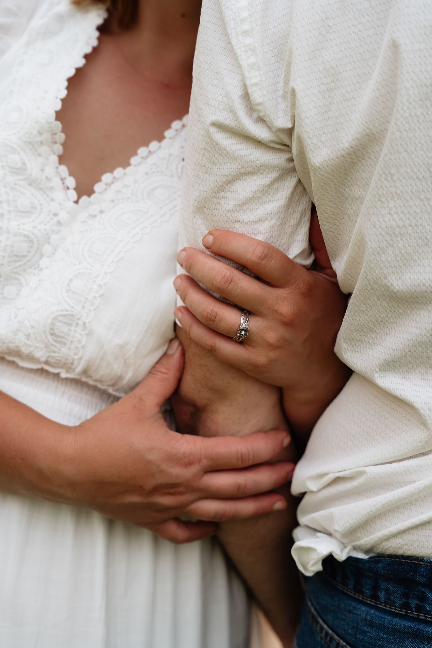 Close-up shot of a couple embracing, showing a white lace dress detail and an engagement ring on hand.