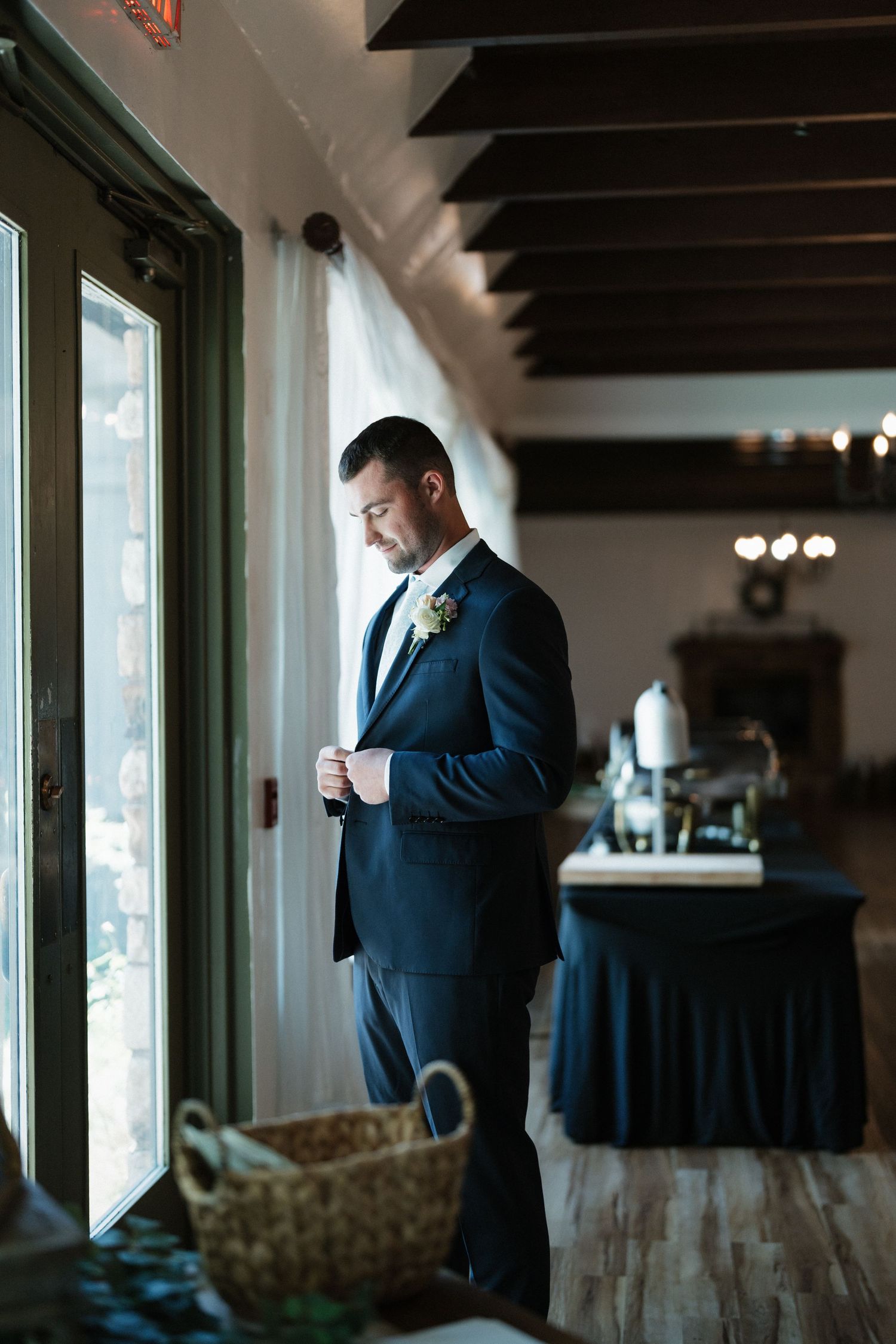 Dapper groom in navy suit adjusting boutonniere near window in elegant indoor venue.