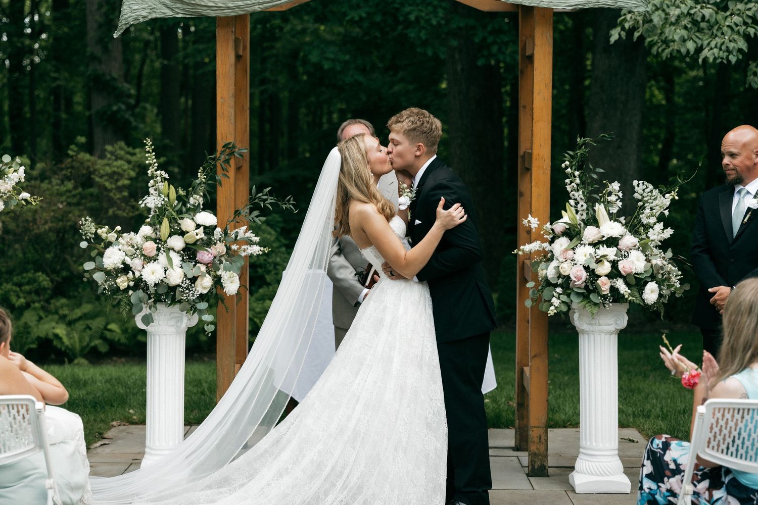 Bride and groom share first kiss at outdoor wedding ceremony with white floral arrangements and wooden arch backdrop.