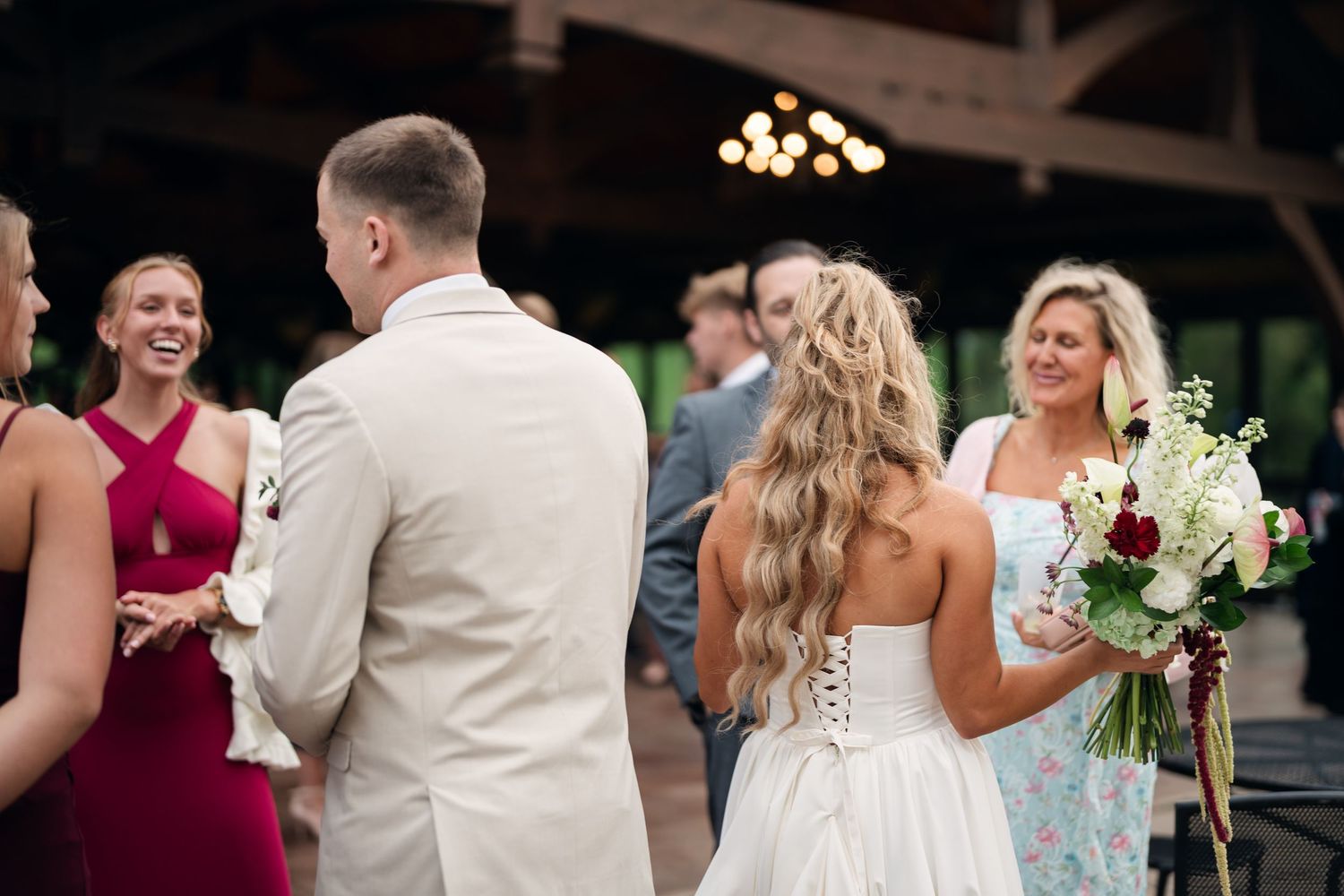 Bride in lace gown holding bouquet smiles with groom in tan suit at outdoor wedding ceremony.