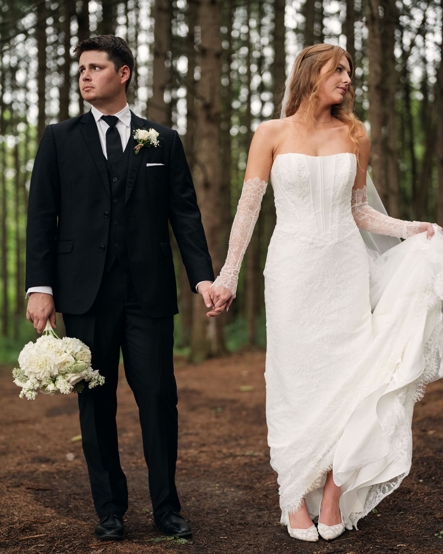 Bride in strapless lace gown with lace gloves and groom in black suit holding hands in pine forest.