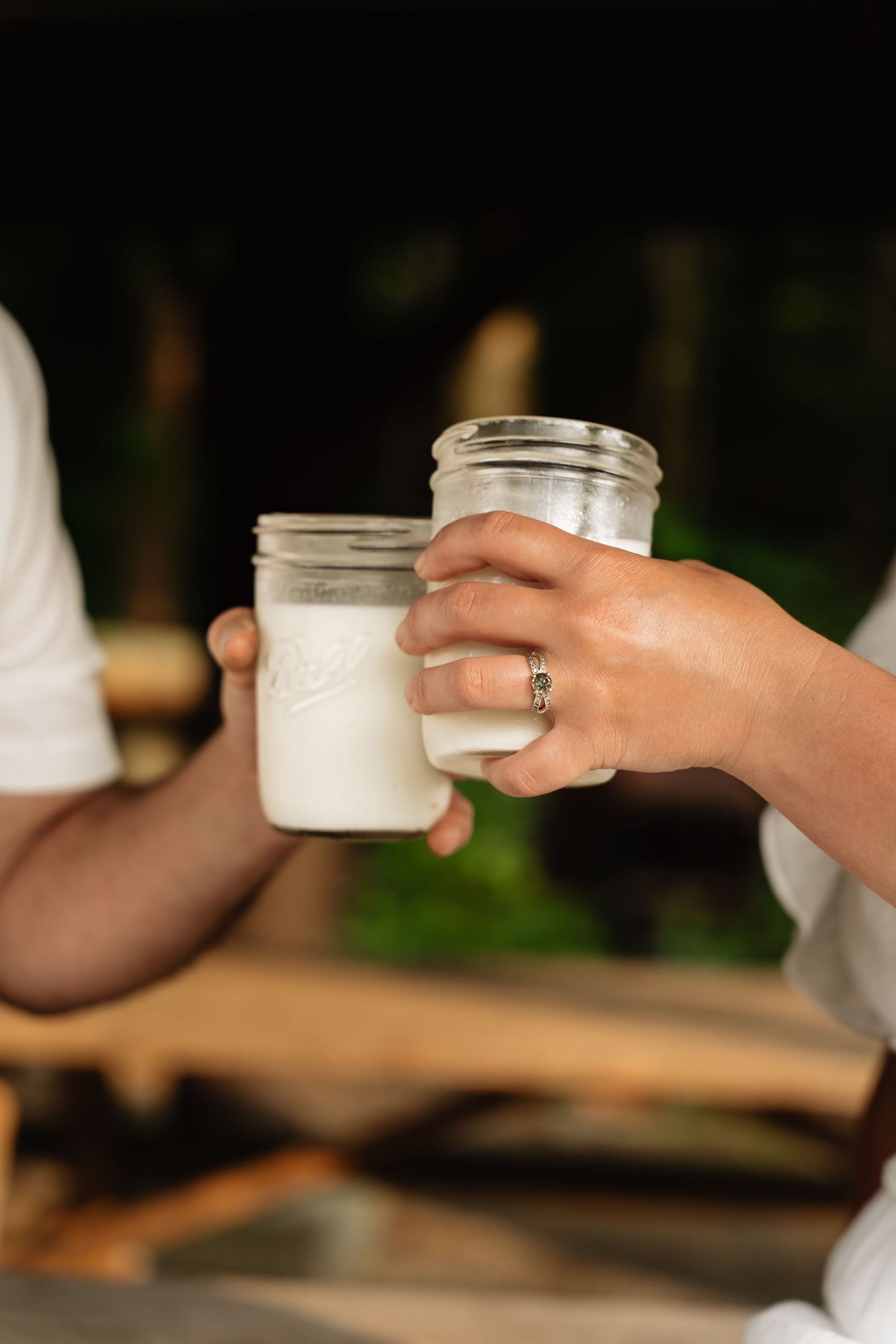 Hands passing glass jars of white cream or lotion on a wooden table with greenery in the background.