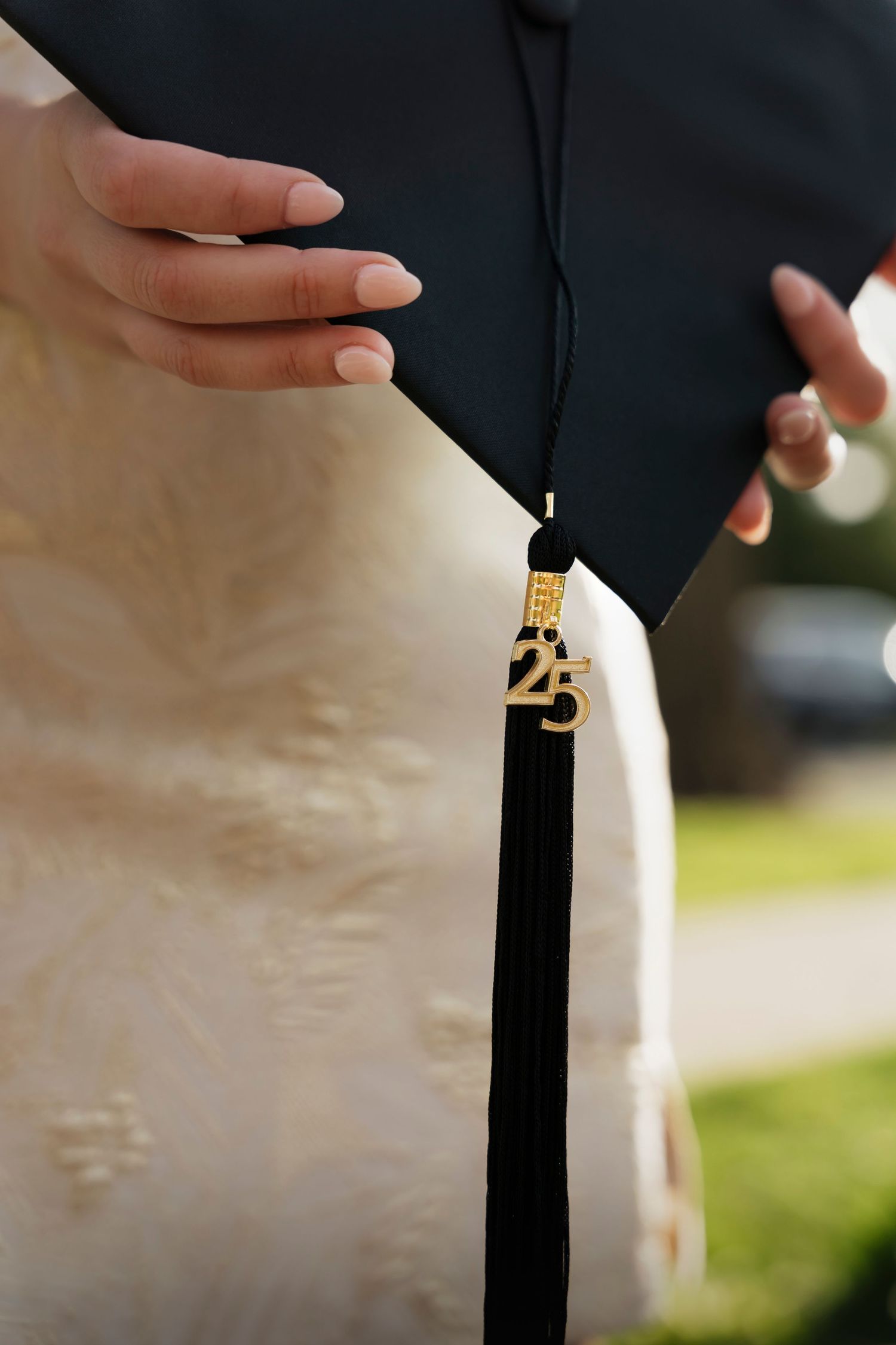 Close-up of a black graduation tassel with gold 2025 charm hanging against white graduation gown in sunlight.