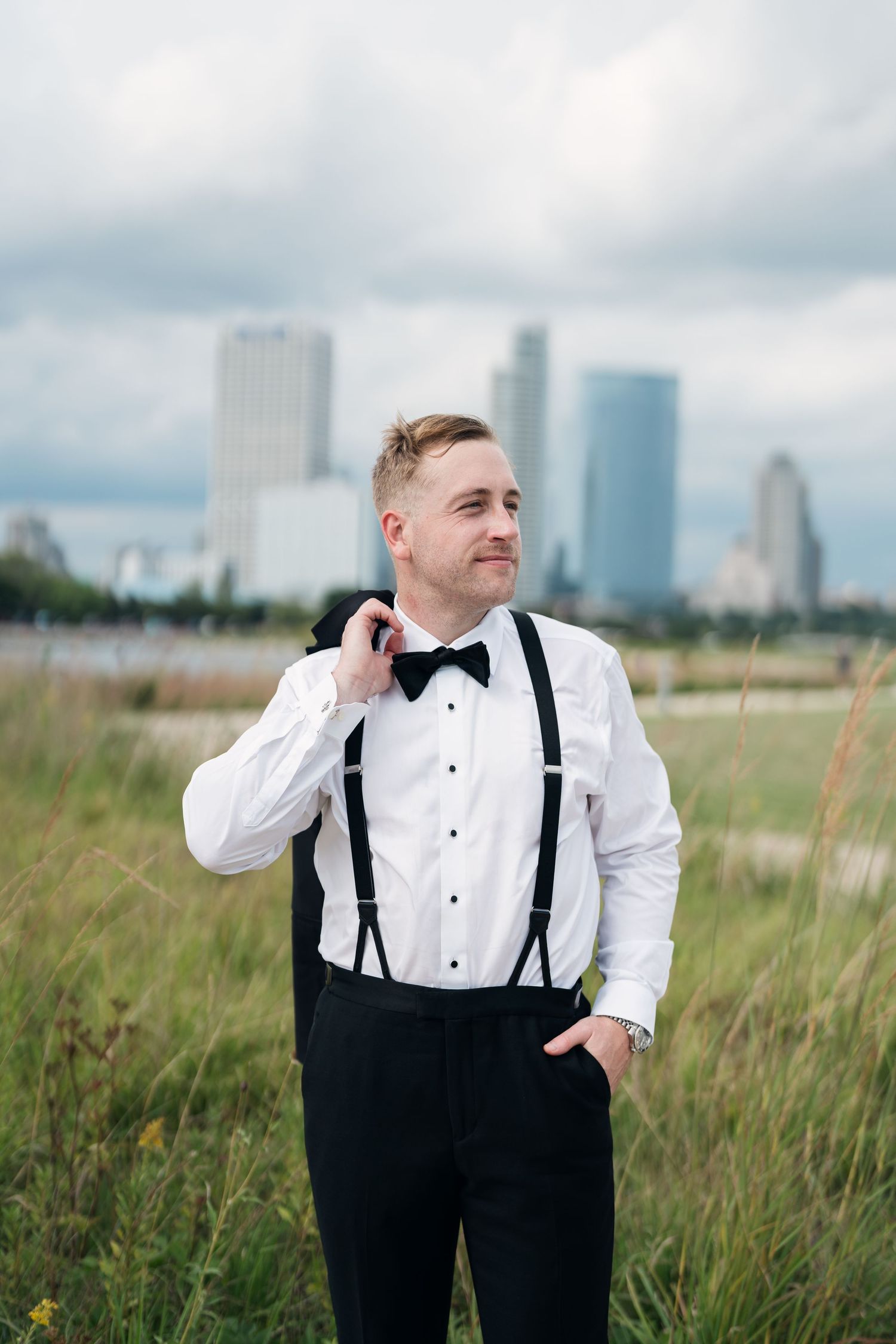 Dapper groom in white tuxedo shirt with black suspenders and bow tie stands in grassy field with city skyline backdrop.