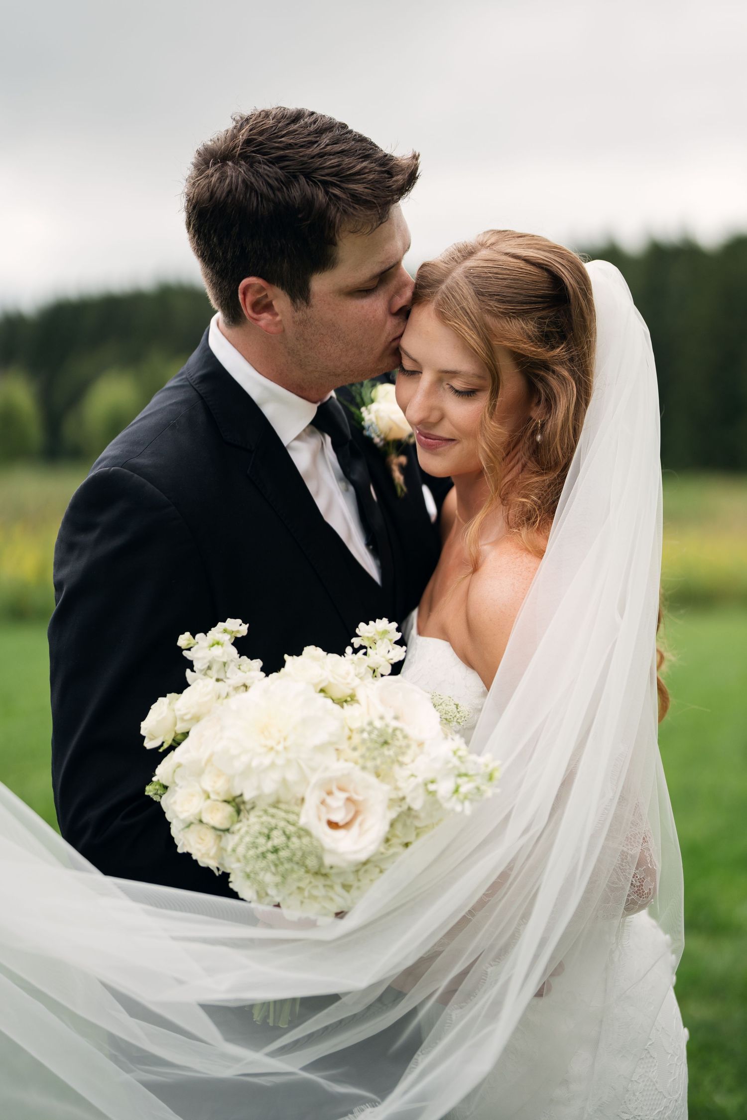 Groom in black tuxedo tenderly kissing bride's temple as her veil flows in the breeze outdoors.