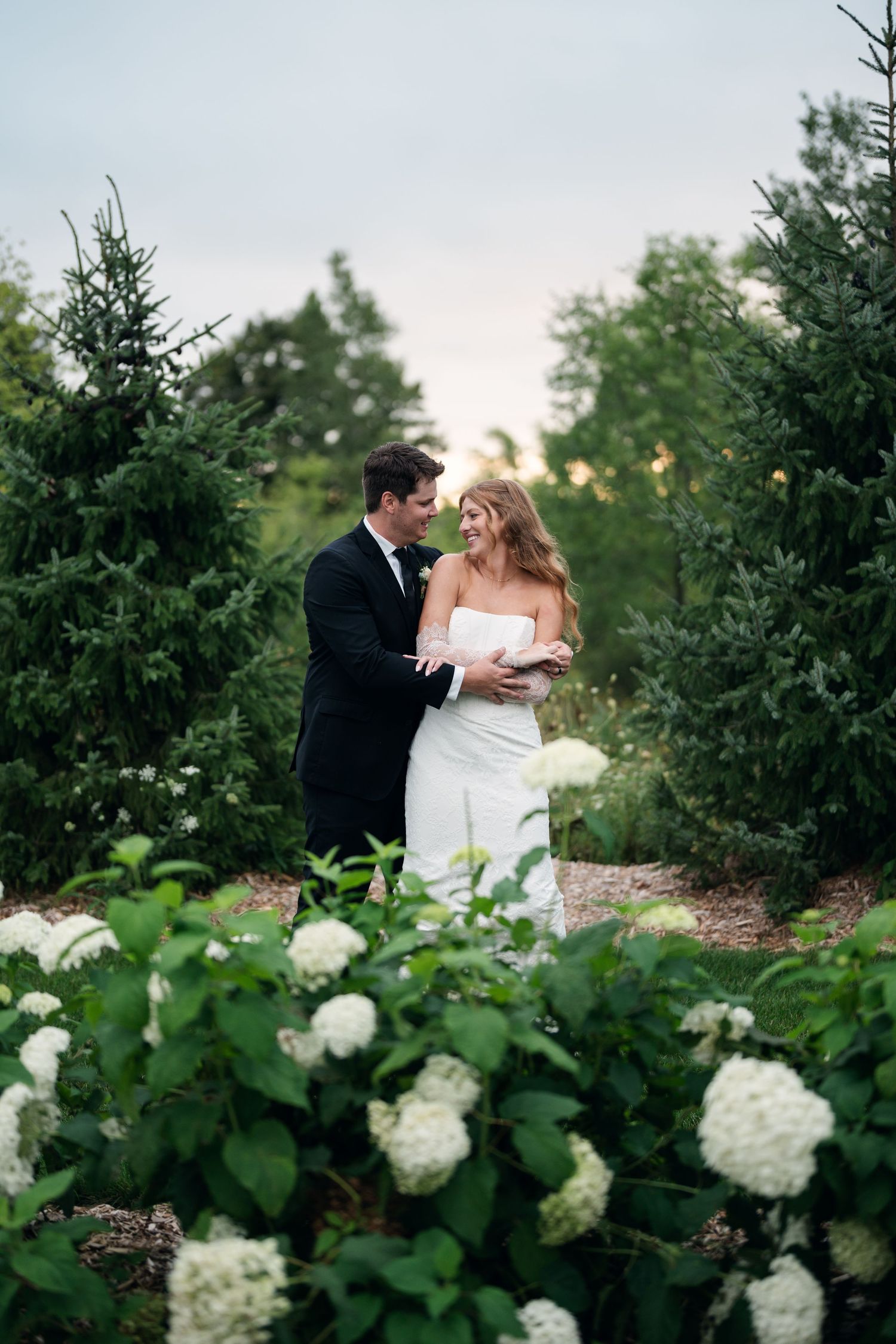Bride and groom share a romantic moment among evergreen trees and white hydrangeas at sunset.
