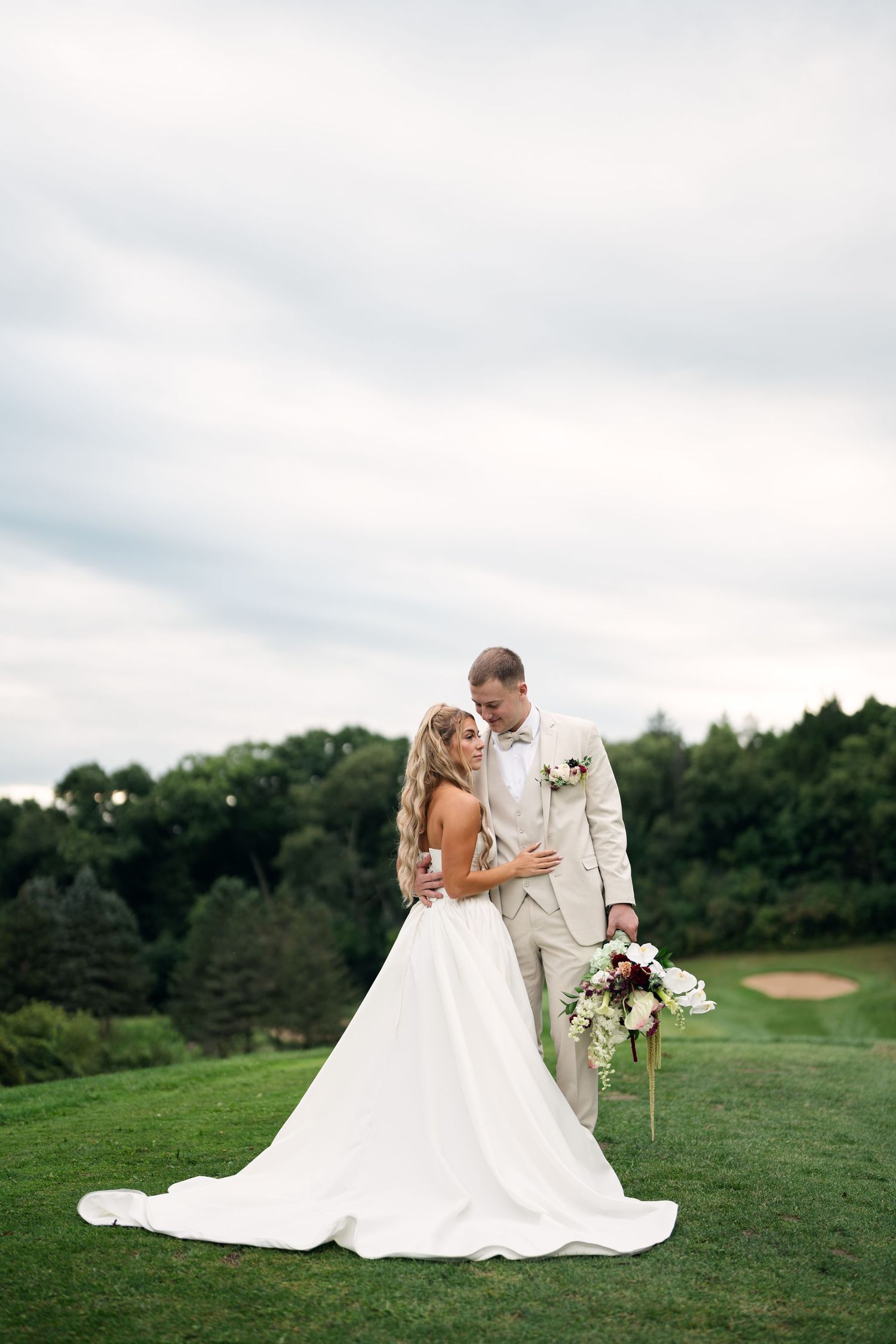Bride and groom embrace on a green golf course, groom holding a bouquet under a dramatic cloudy sky.