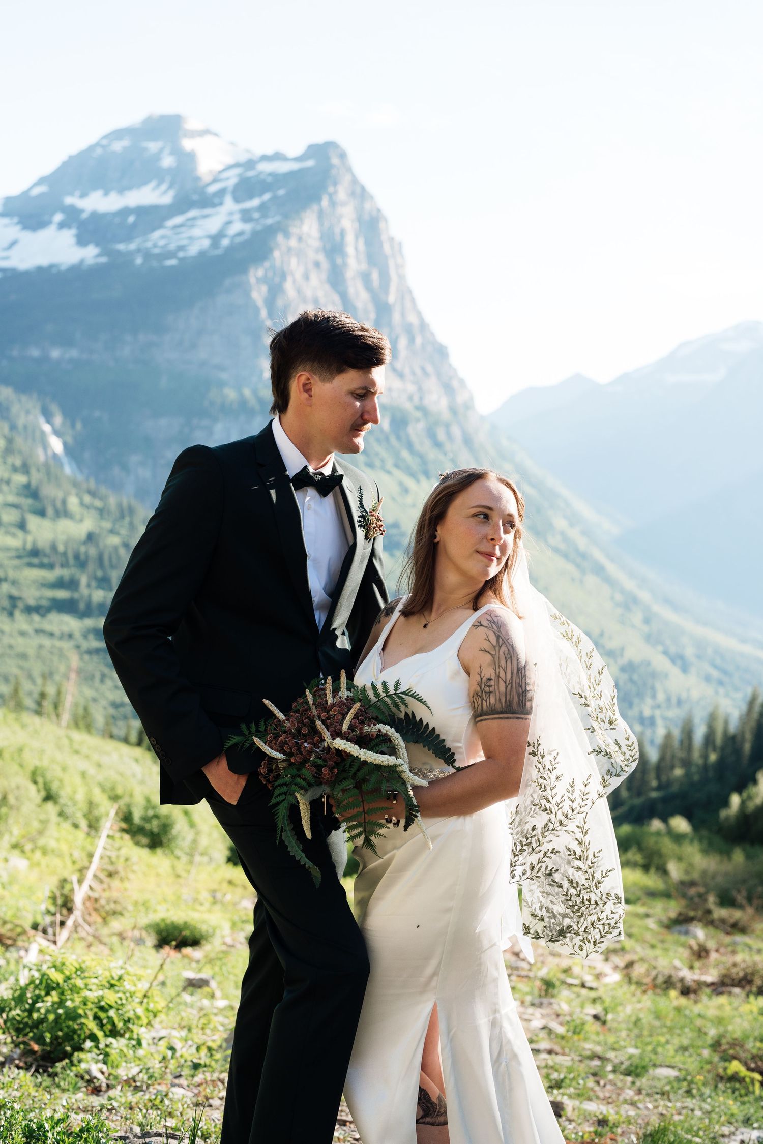 A bride and groom pose together in wedding attire with stunning mountain scenery in Glacier National Park.