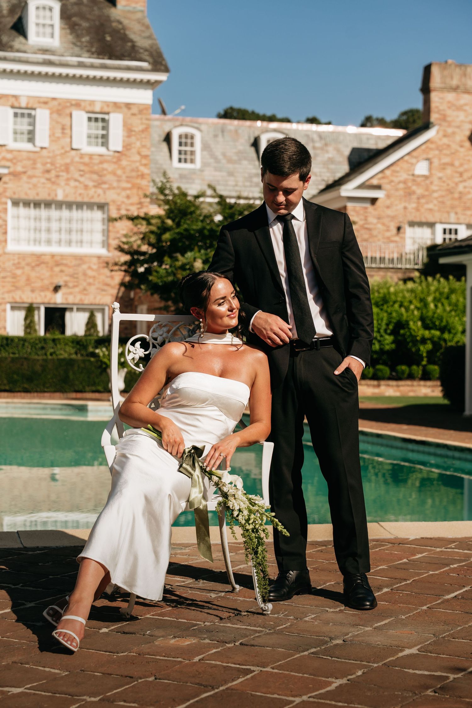 Wedding couple poses playfully on brick patio next to swimming pool at elegant brick estate venue.