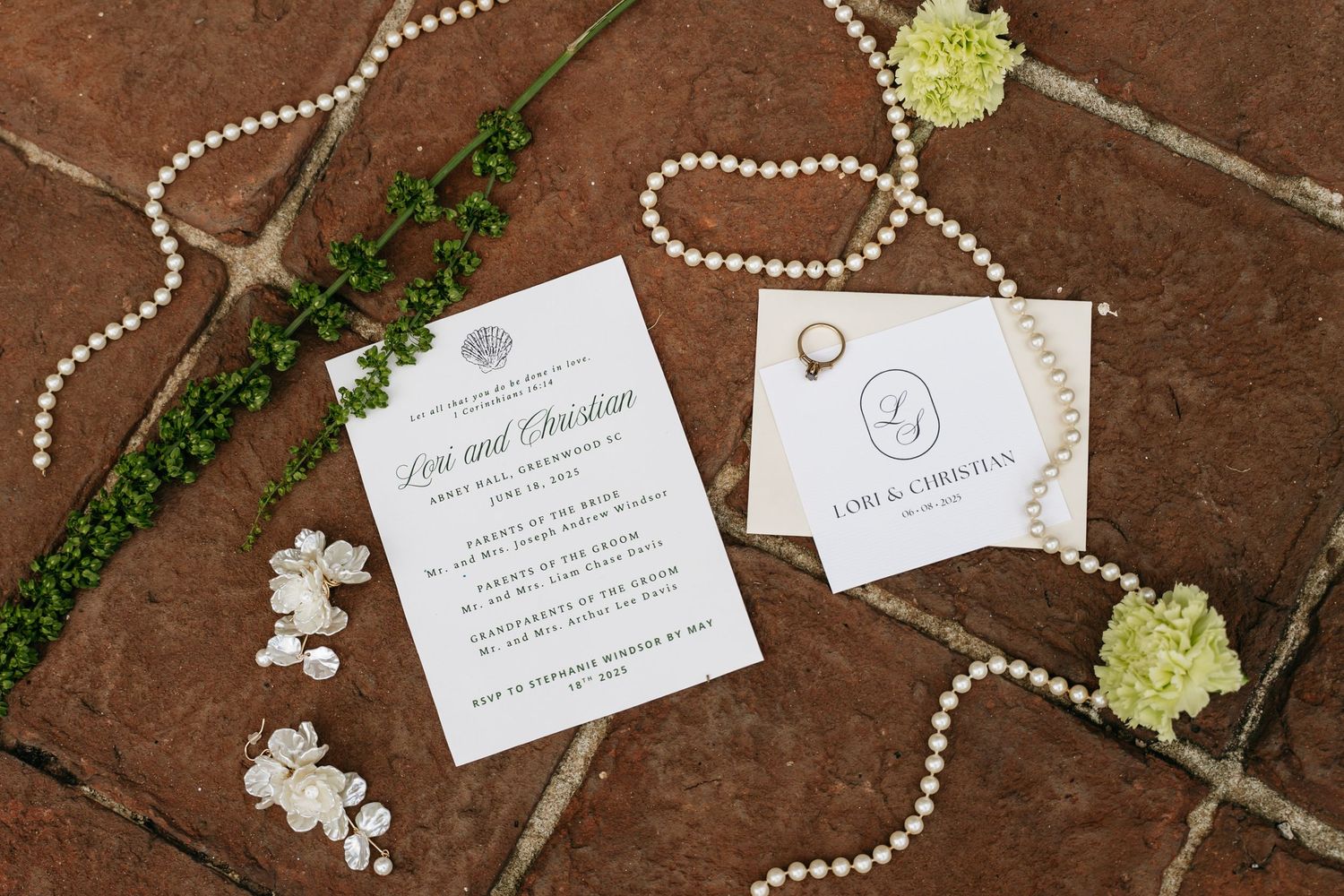 Elegant wedding invitations and pearl necklaces arranged on a rustic wooden surface with delicate green floral accents.