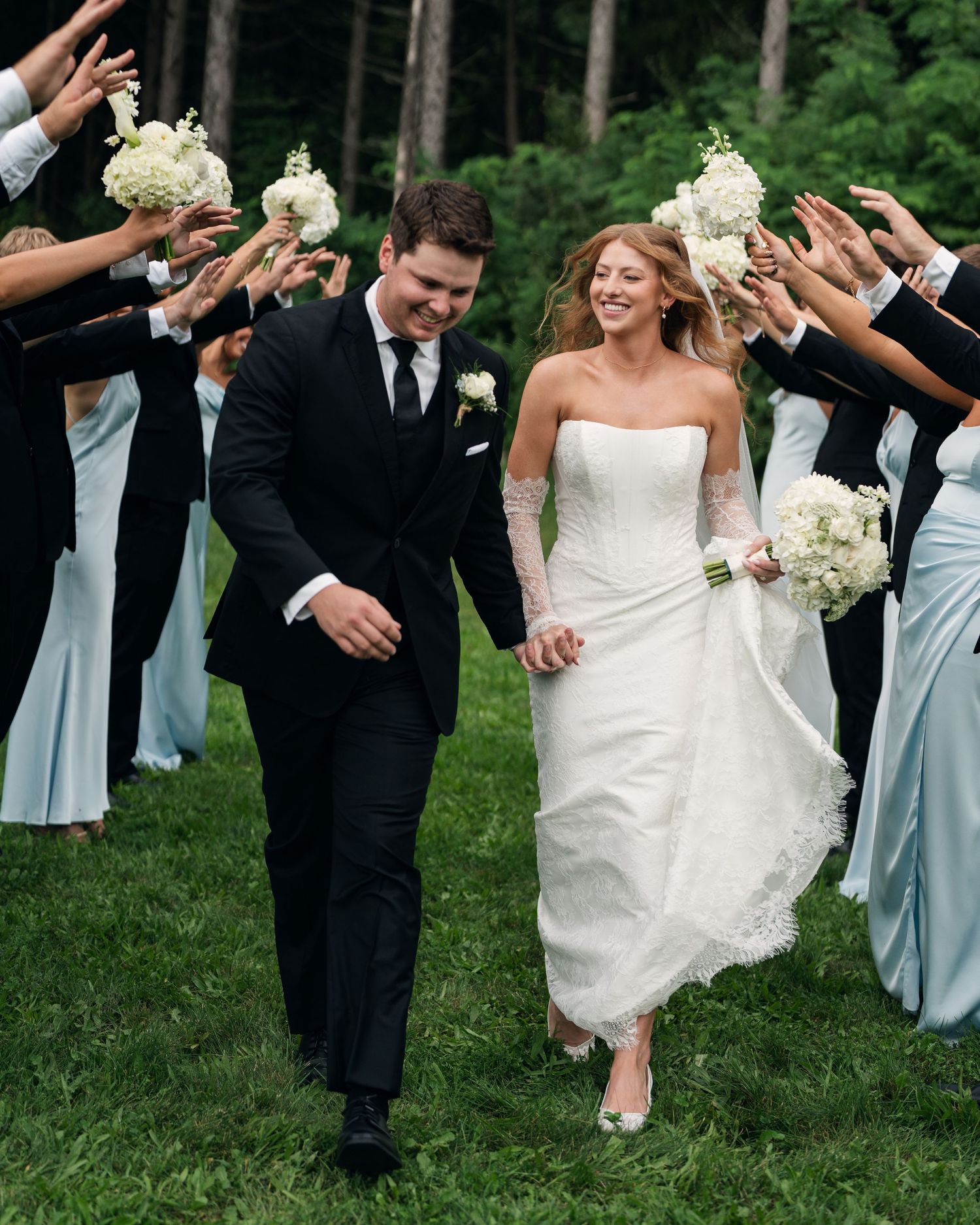 Smiling newlyweds walk through a tunnel of raised hands held by wedding party members in black and blue attire.