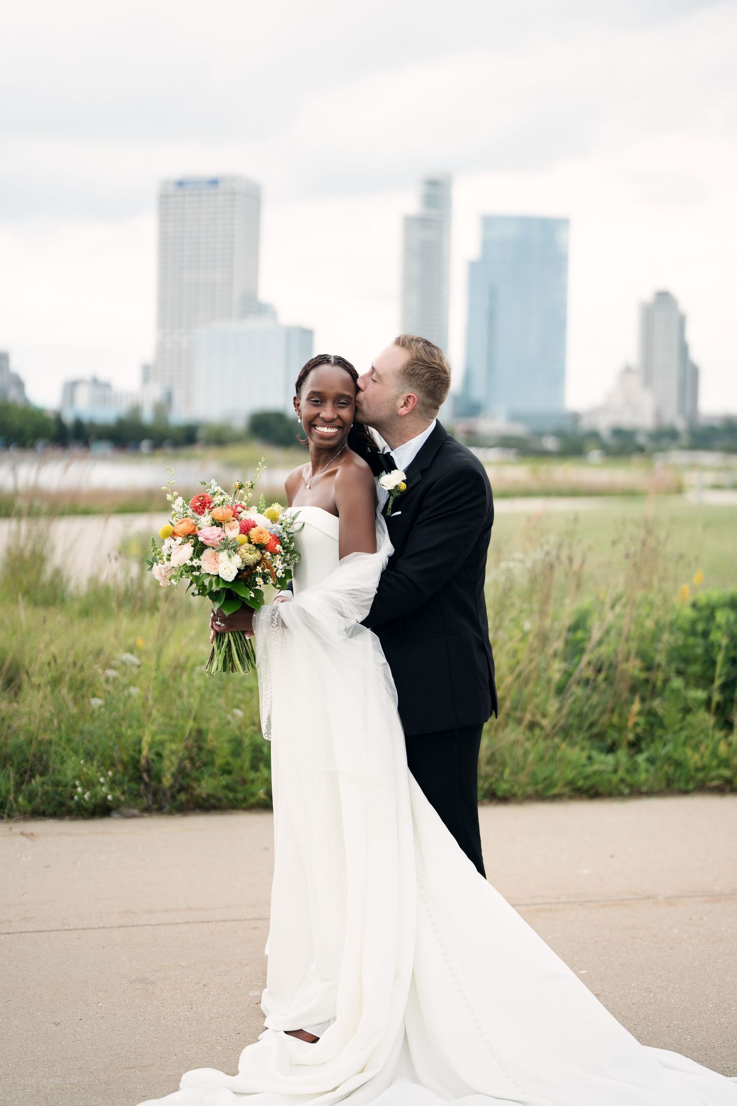 Bride and groom share a romantic kiss with city skyline backdrop, bride holding colorful bouquet in white gown.