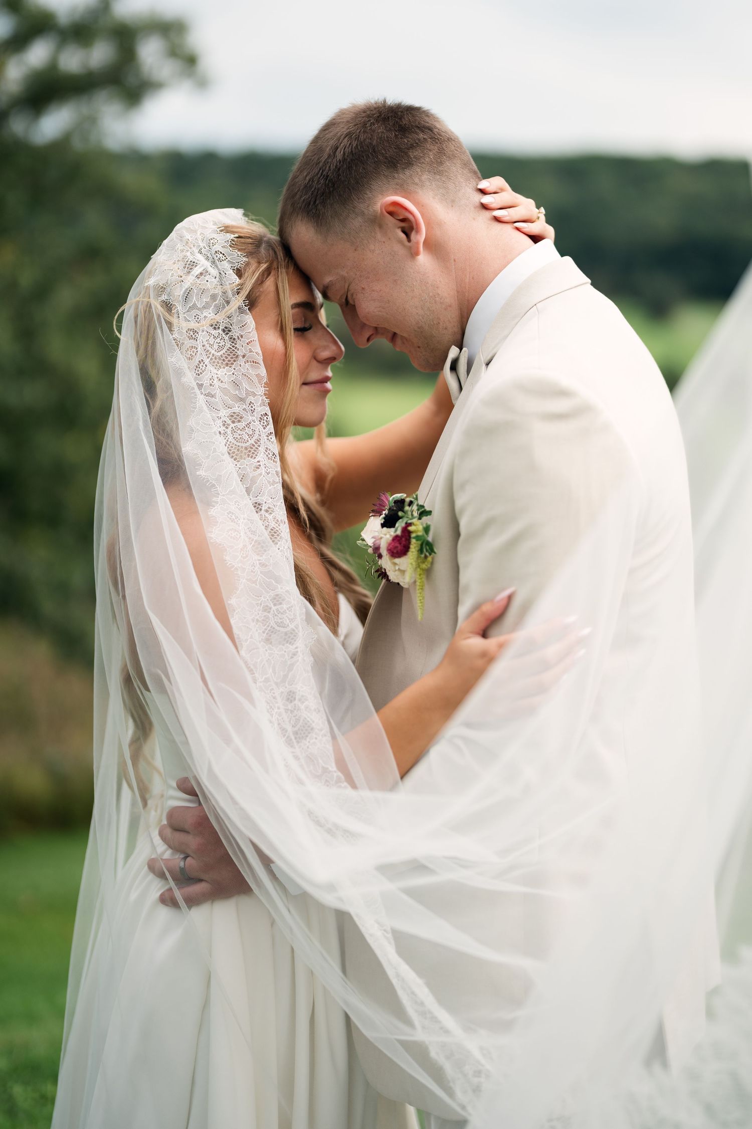 Bride and groom share an intimate forehead-to-forehead moment wrapped in a flowing veil with green fields behind.