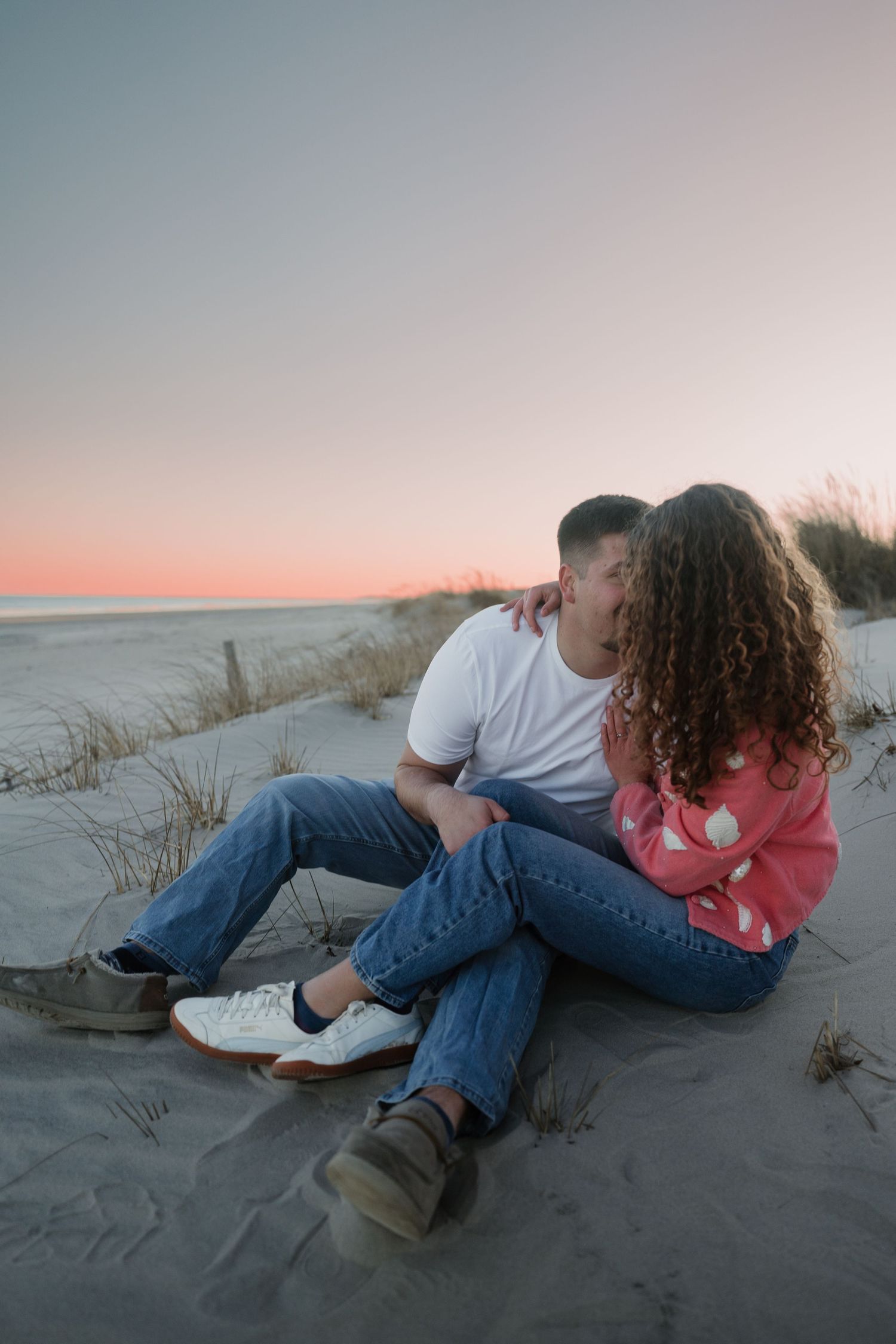 Couple relaxes on sandy beach dunes during sunset engagement photo session.