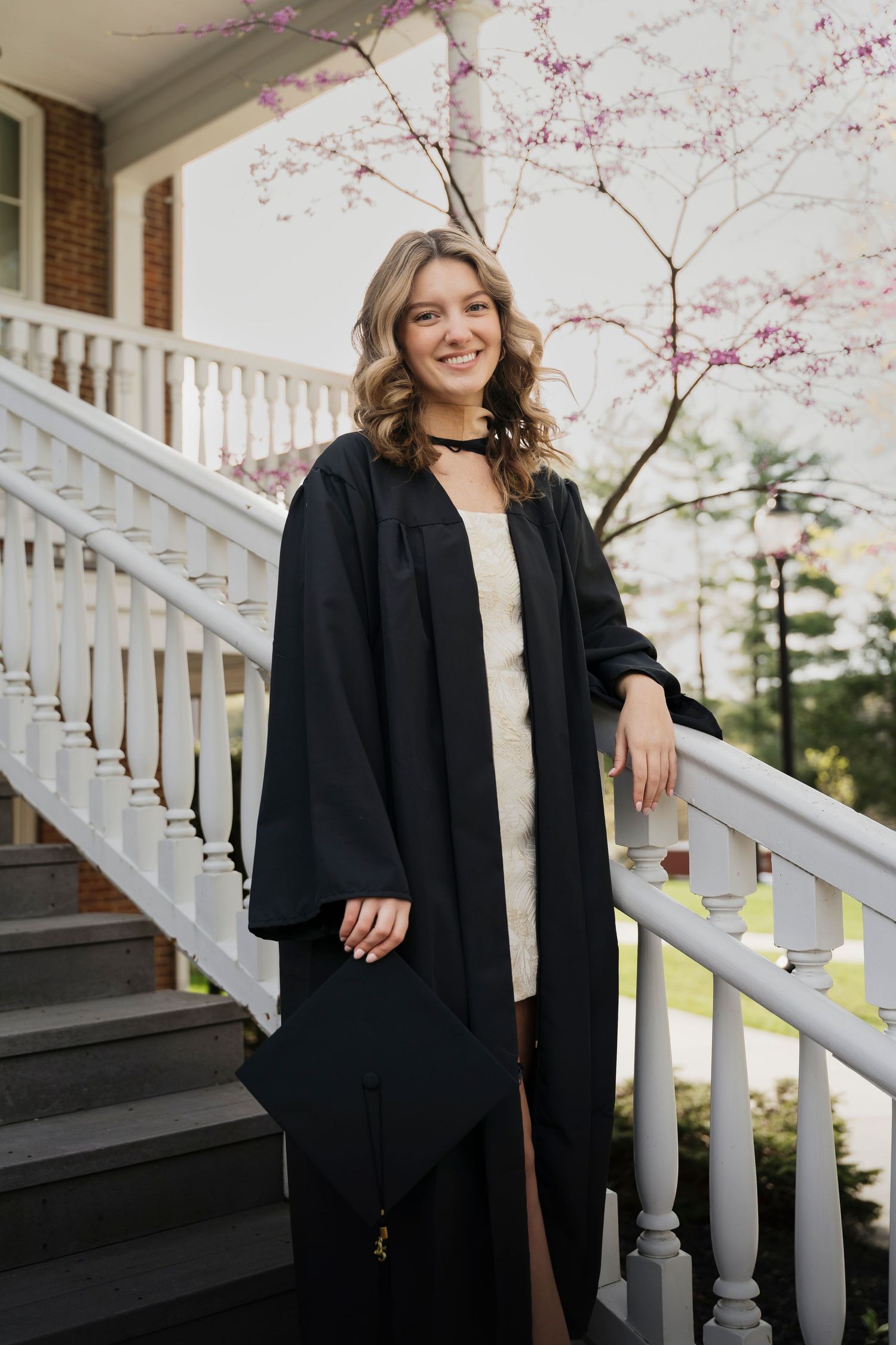 Graduate poses by white porch railing with pink cherry blossoms in the background.