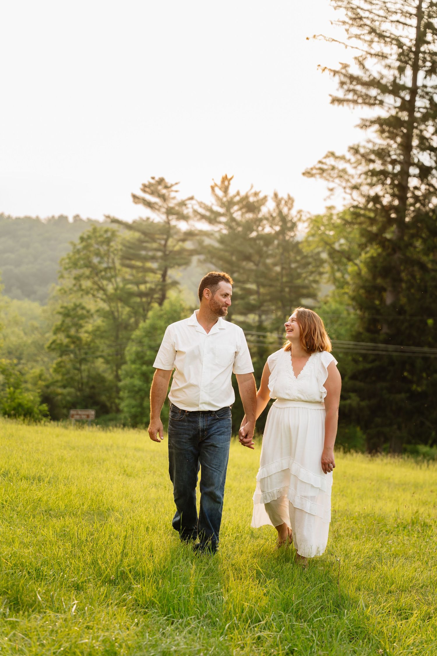 A couple holds hands while walking through a sunlit meadow with pine trees and rolling hills in the background.