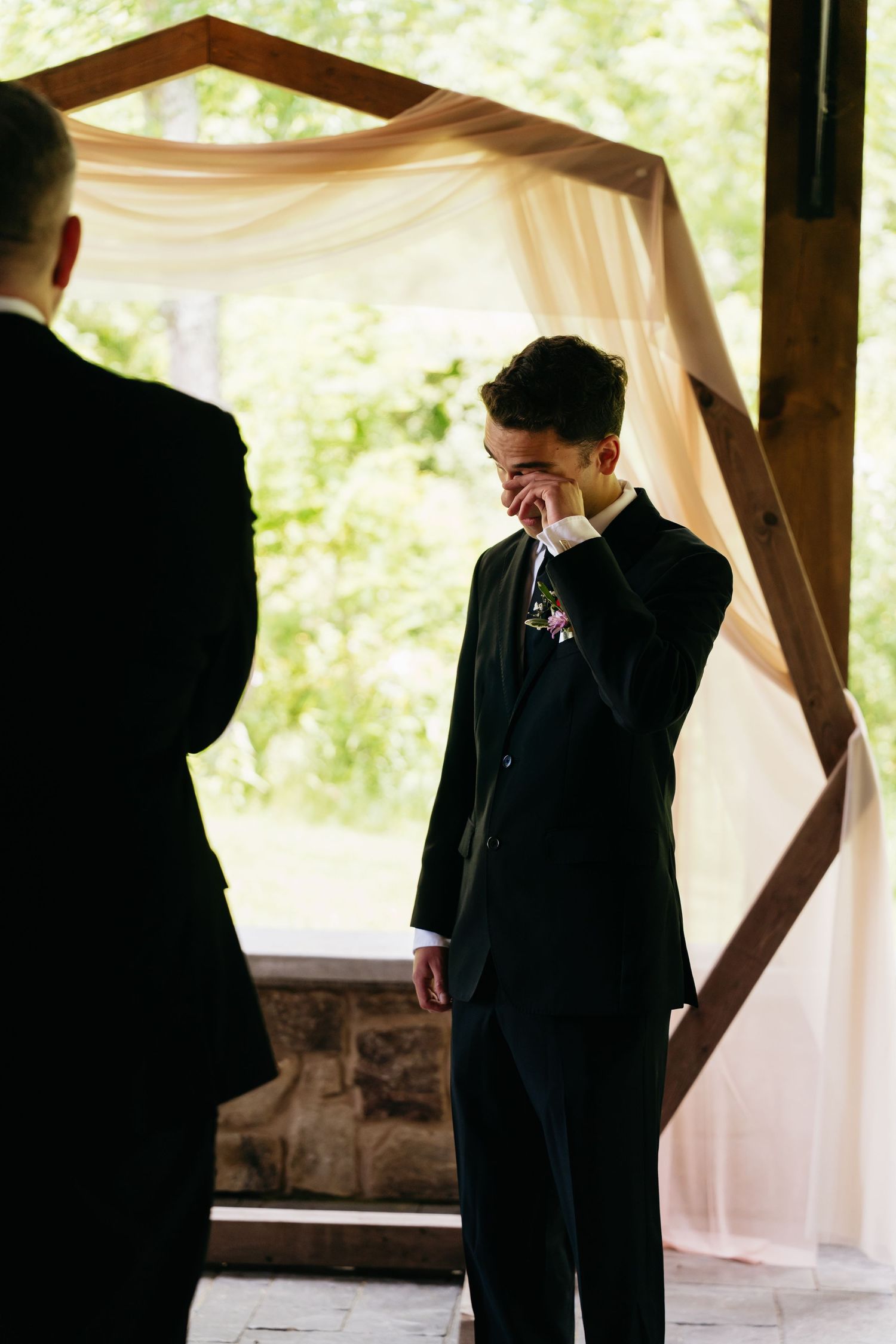 An emotional moment at a wedding ceremony as someone in a black suit wipes tears beneath a draped fabric arch.