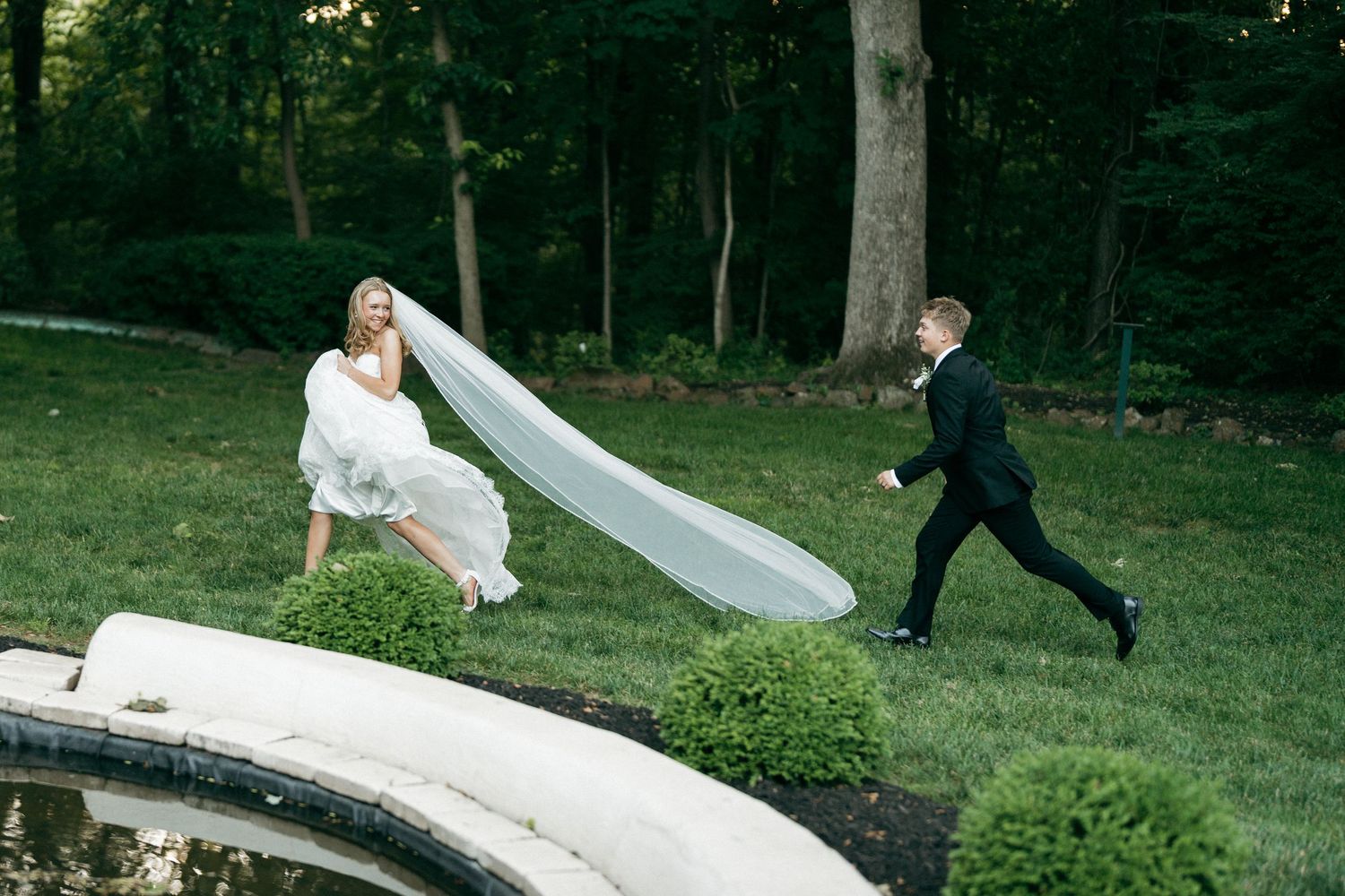Bride in white gown and veil runs with groom in tuxedo across lush green garden near ornate fountain.