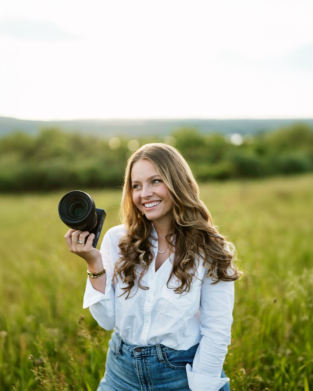 Smiling woman holding a camera in a sunny green field, wearing a white shirt and jeans, with trees in the background.