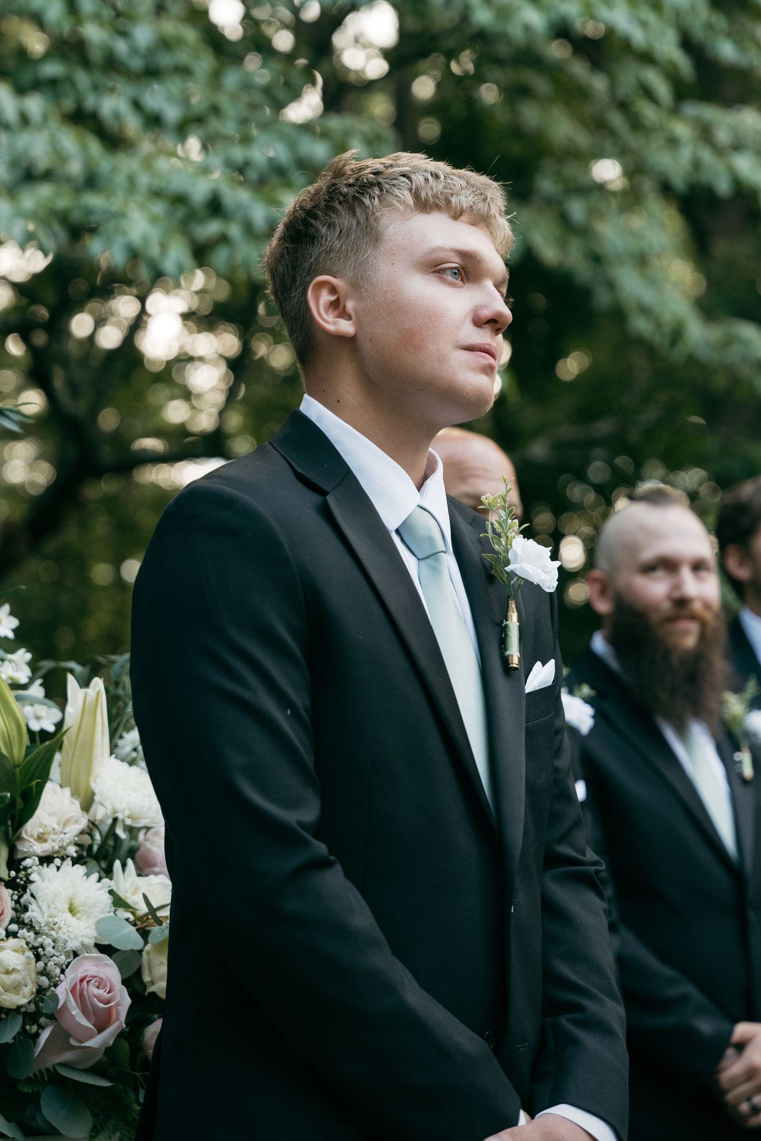 Groom in black suit with white boutonniere stands at outdoor wedding ceremony among lush green trees.
