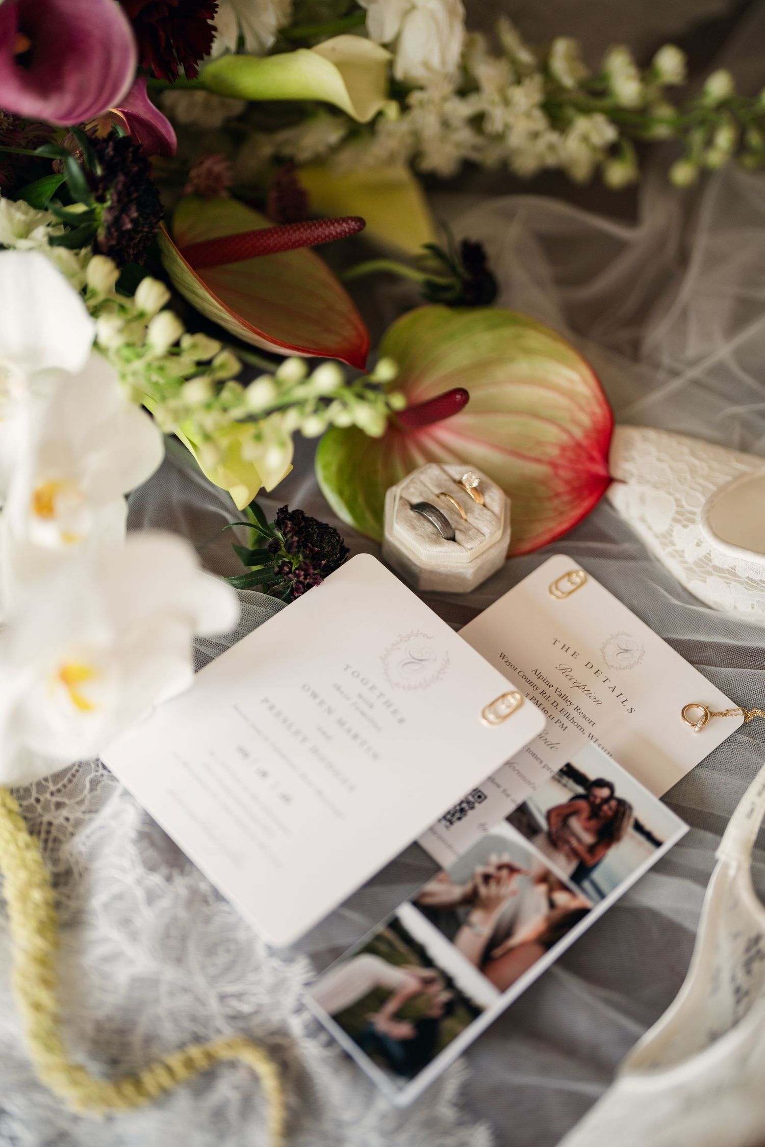 Wedding flat lay featuring rings, floral bouquet, invitation suite, and photo strip on elegant gray fabric.