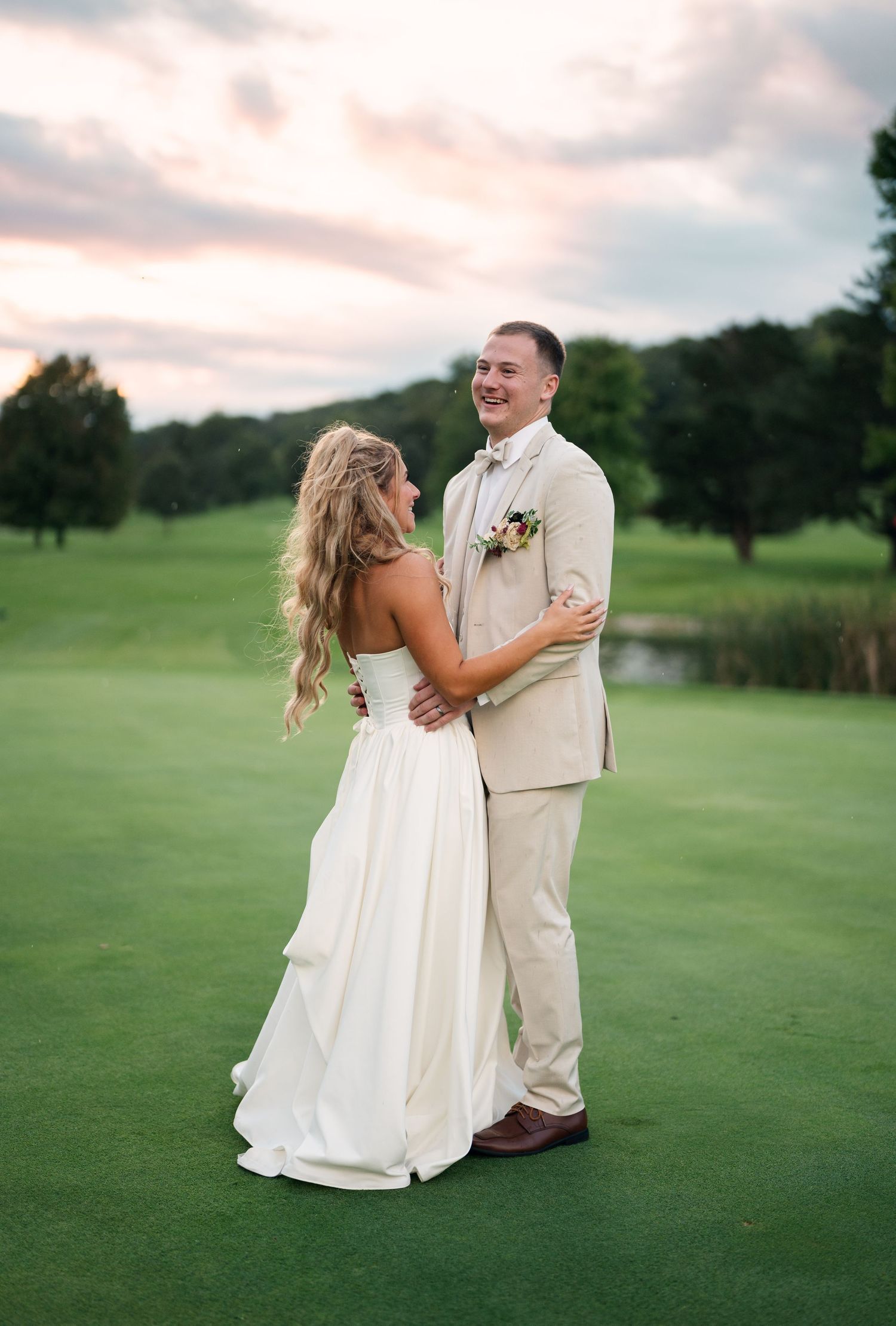 Bride and groom laugh together on a golf course at sunset, with a scenic pond and trees in the background.