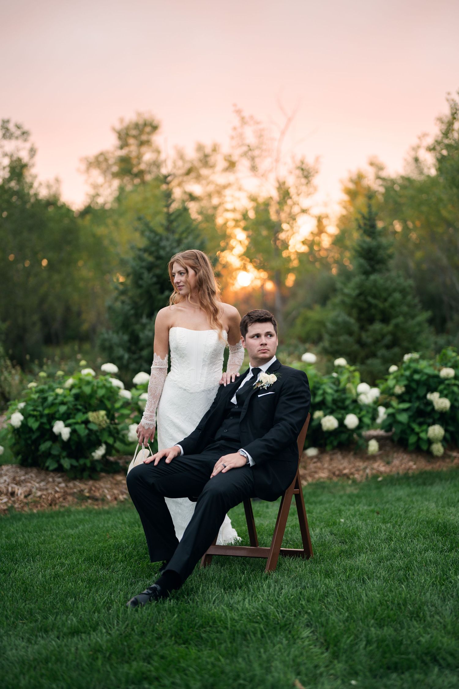 Bride in white gown stands behind groom in black tuxedo seated on wooden chair at sunset garden wedding.