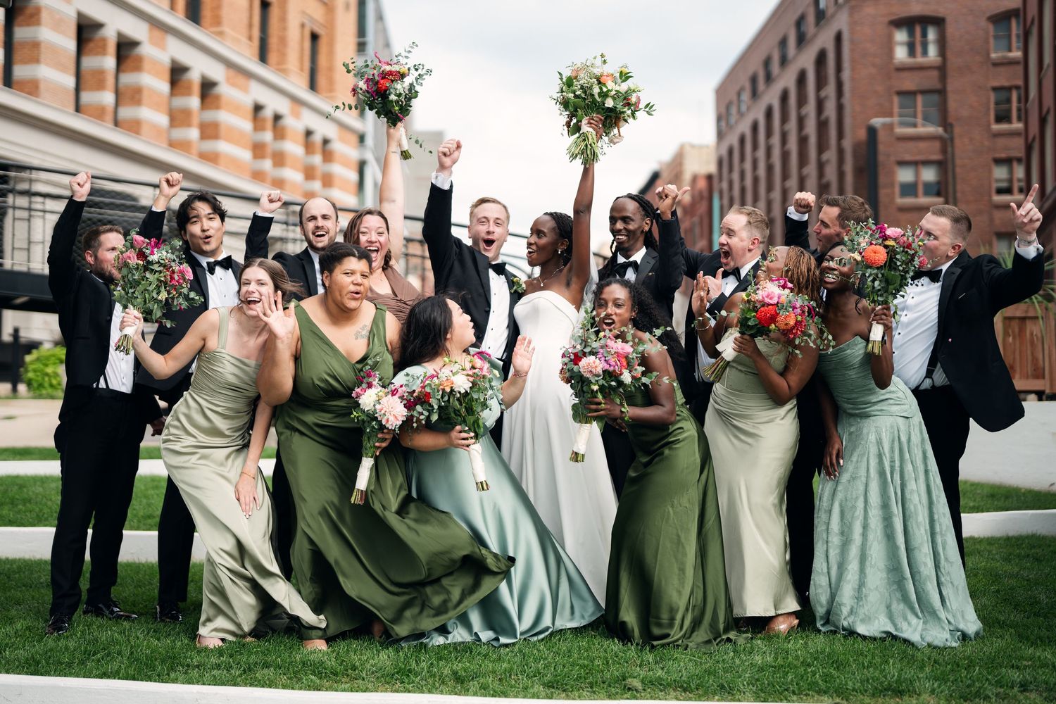 Diverse wedding party celebrating outdoors in green bridesmaid dresses and black tuxedos.