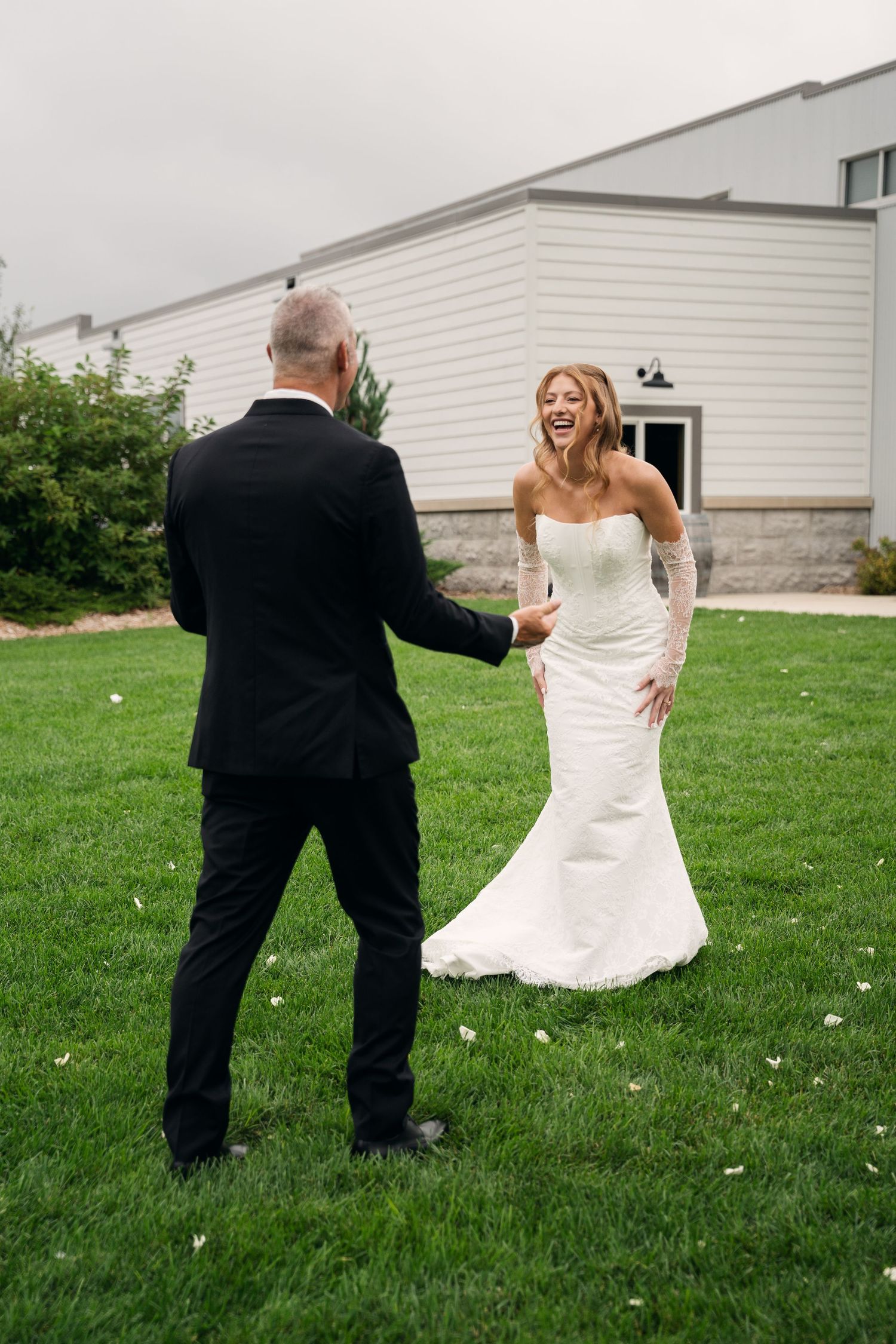 Laughing bride in strapless white gown shares a joyful first look moment with her groom on green lawn.