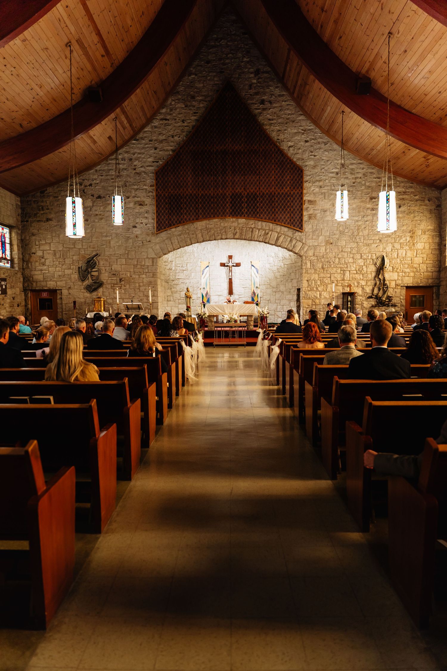 Interior view of a modern church sanctuary with stone walls, wooden pews, and hanging pendant lights along the aisle.