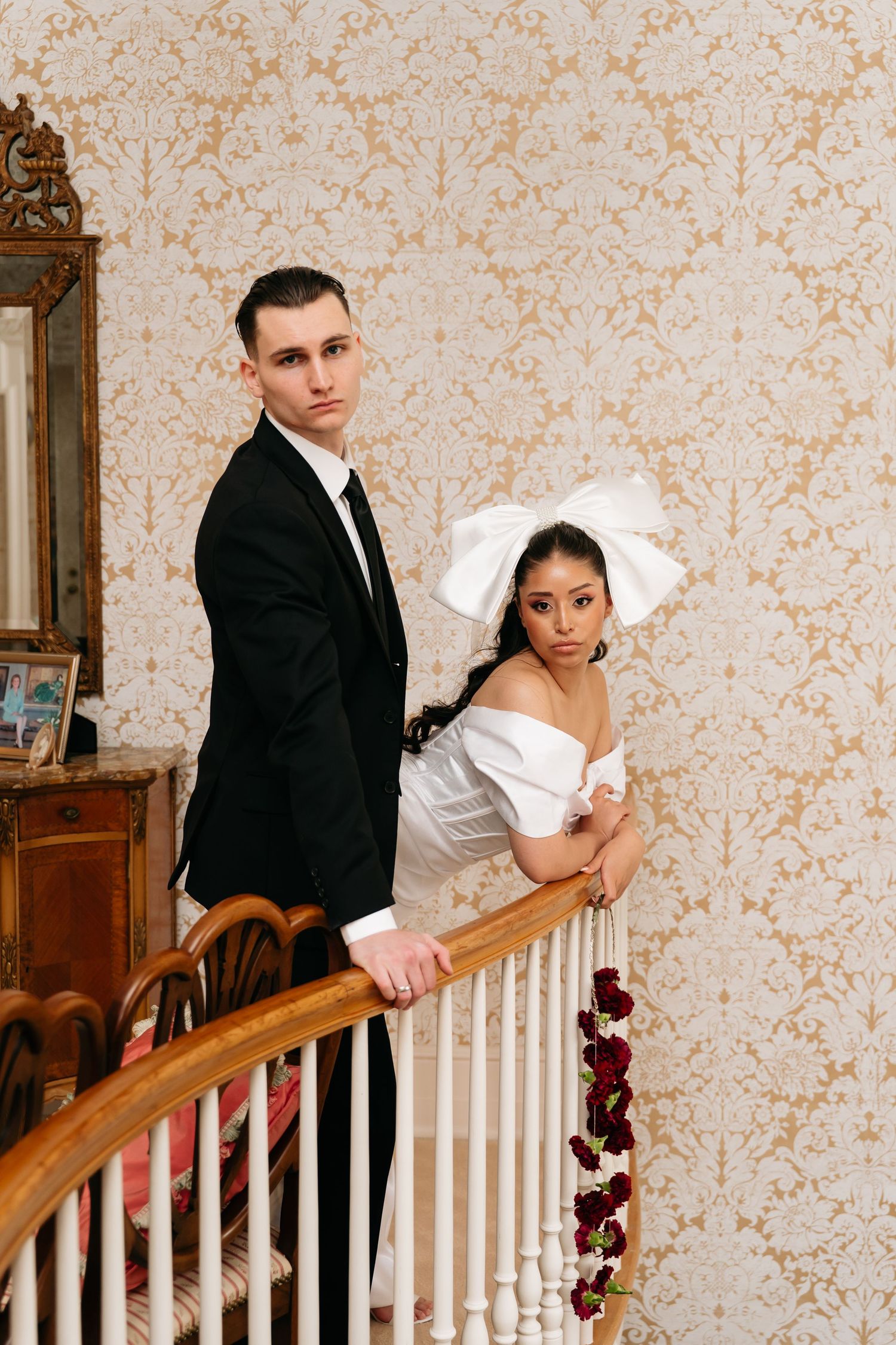 Elegant couple in wedding attire posing on a staircase with floral decor and vintage wallpaper backdrop.
