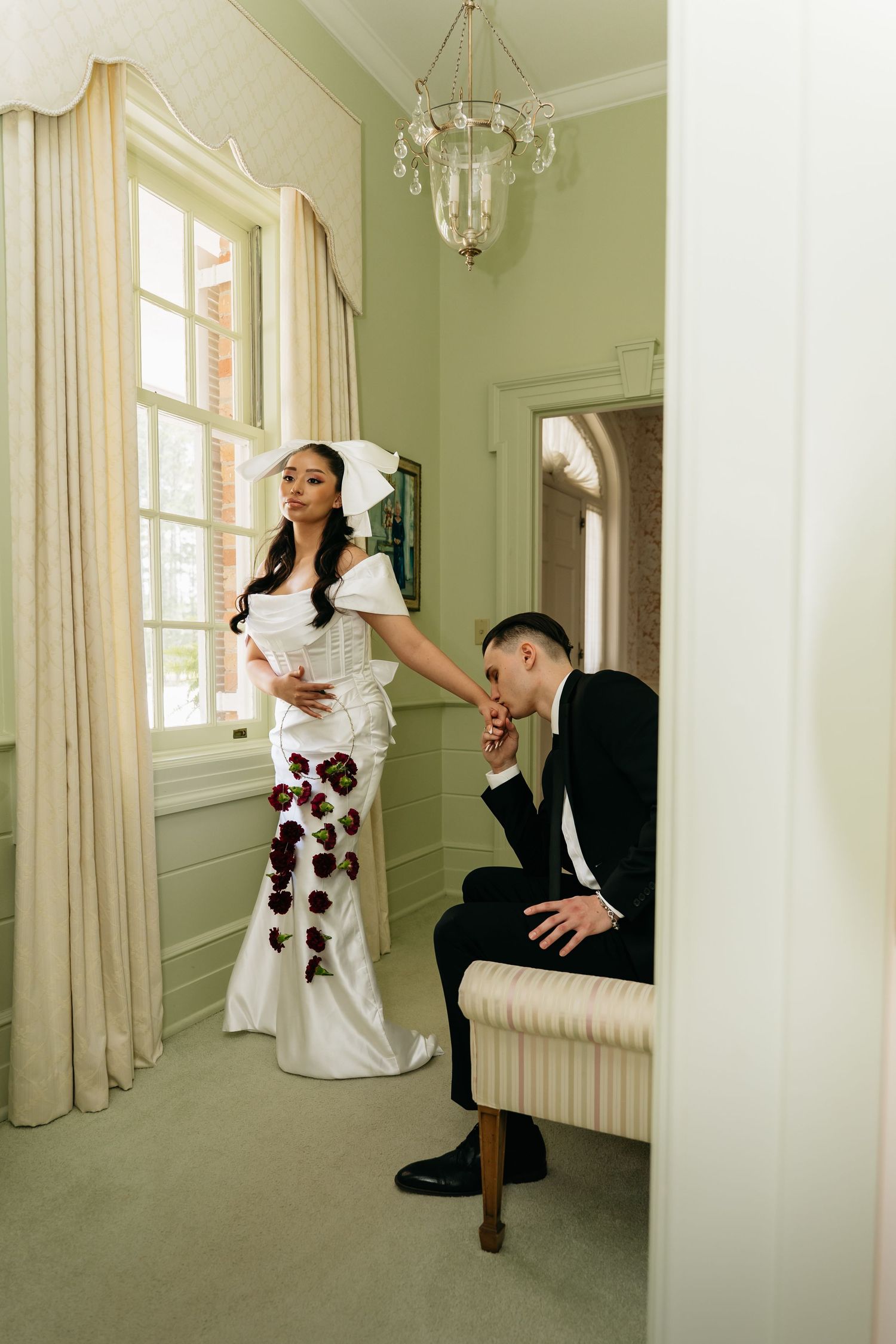 Bride in white floral gown with bow headpiece stands by window as groom kisses her hand in elegant green room.