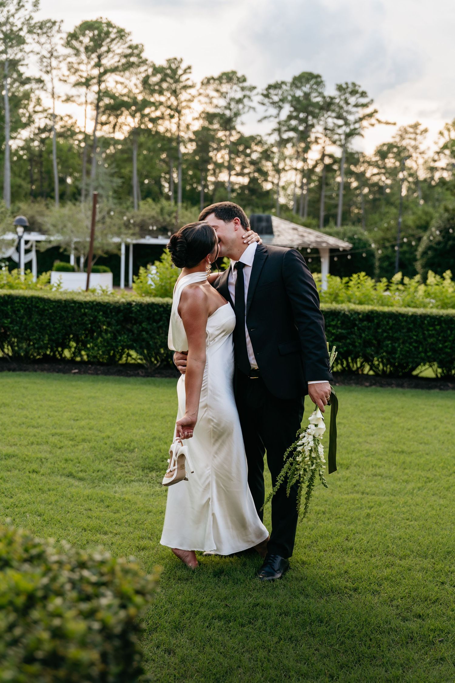 A couple shares a romantic moment on a lush green lawn at sunset with tall trees in the background.
