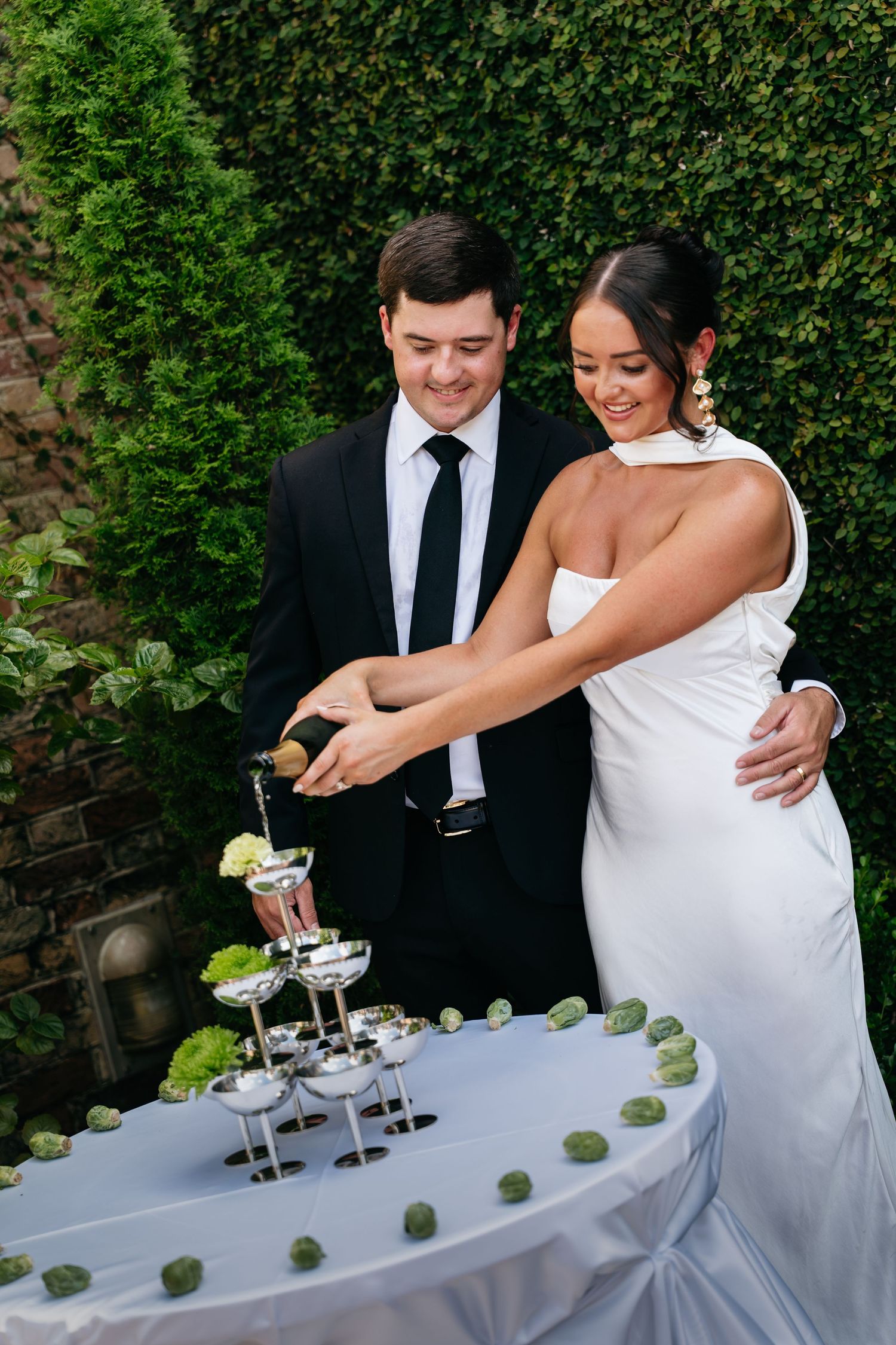 A couple in wedding attire cuts their wedding cake together at an outdoor reception with greenery in the background.