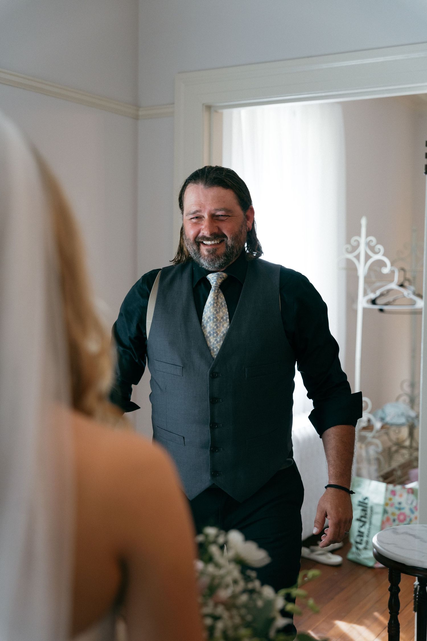 Smiling bearded man in dark vest and floral tie reacts joyfully upon seeing bride during wedding preparations.