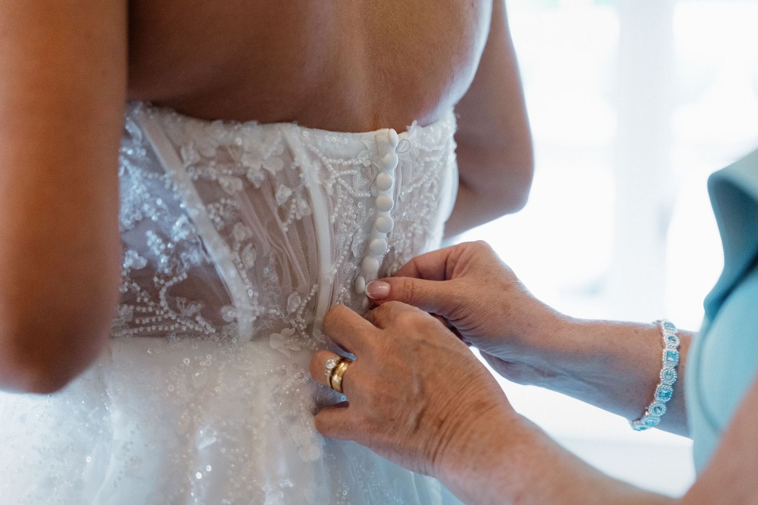 Hands fastening delicate buttons on the back of a lace wedding dress during bridal preparations.
