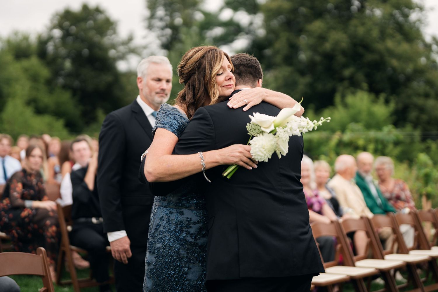 A woman in a blue dress shares an emotional hug with a groom during an outdoor wedding ceremony.