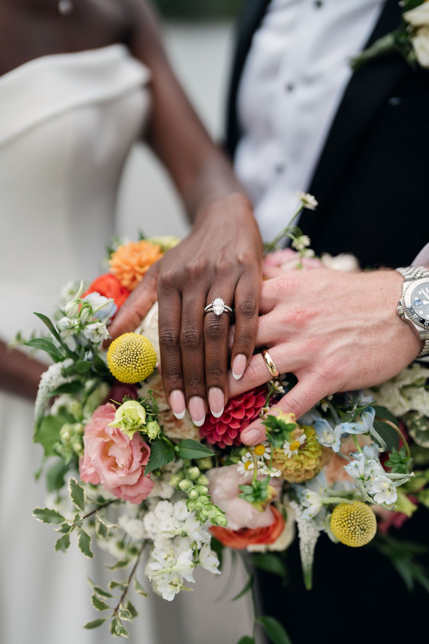 Close-up of interracial newlyweds displaying wedding rings on vibrant bridal bouquet with roses and yellow blooms.