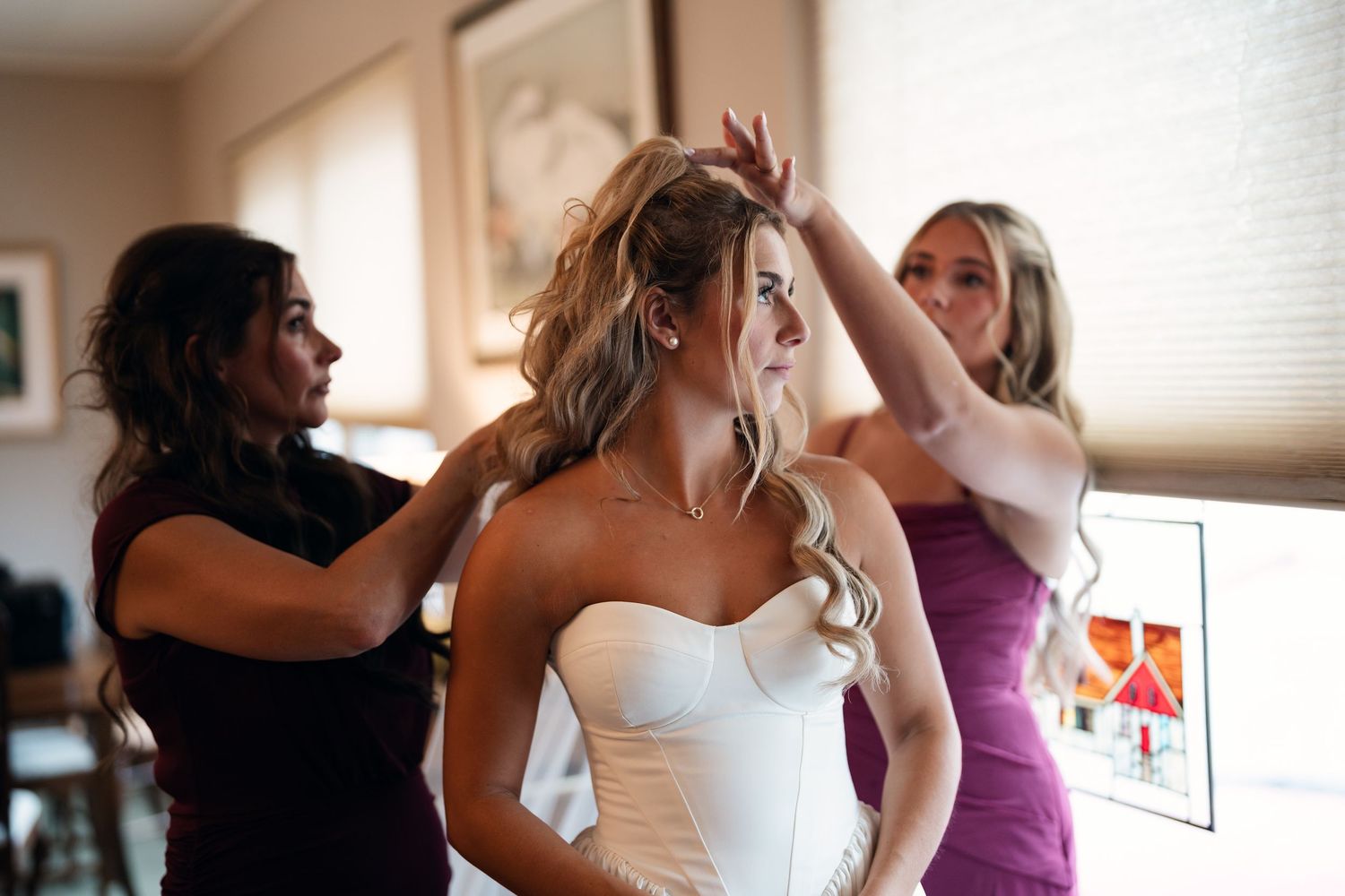 Bride in white strapless gown gets hair styled by two bridesmaids before wedding ceremony.