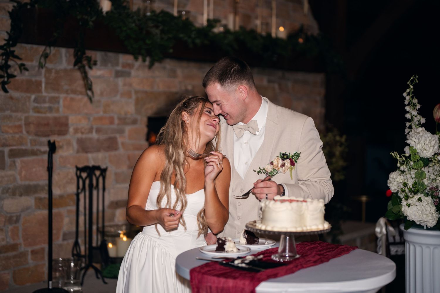 Bride and groom share a kiss while cutting their white wedding cake at a romantic rustic venue reception.