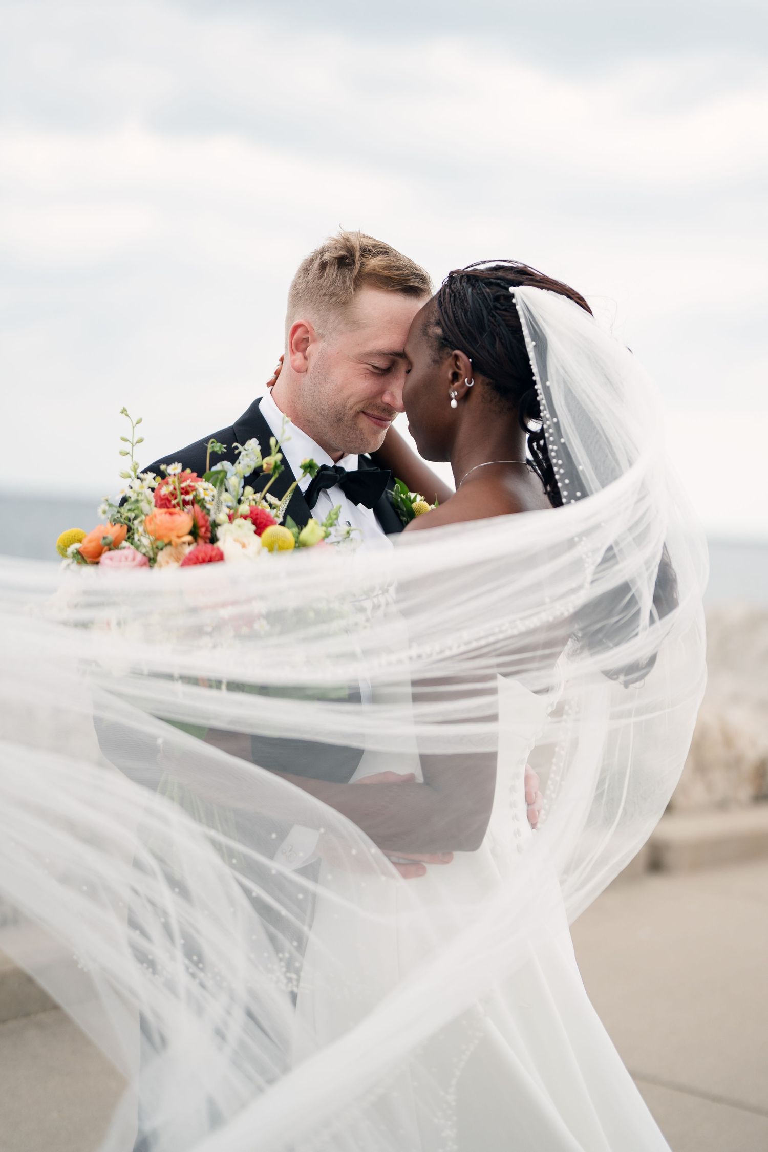 Interracial couple sharing romantic moment with flowing veil on wedding day near waterfront.