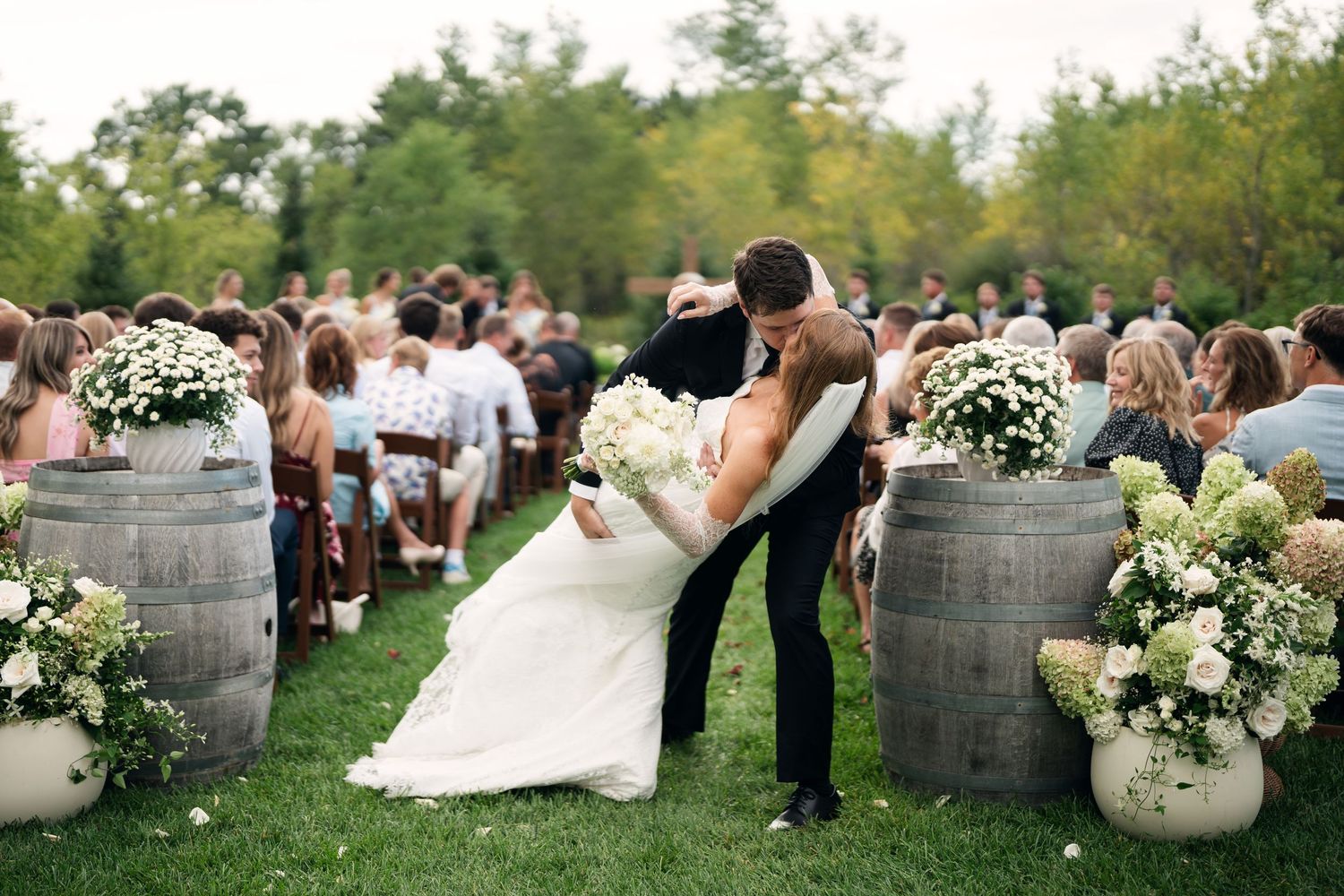 Bride and groom sharing a romantic dip kiss at outdoor wedding ceremony lined with barrel planters and white flowers.