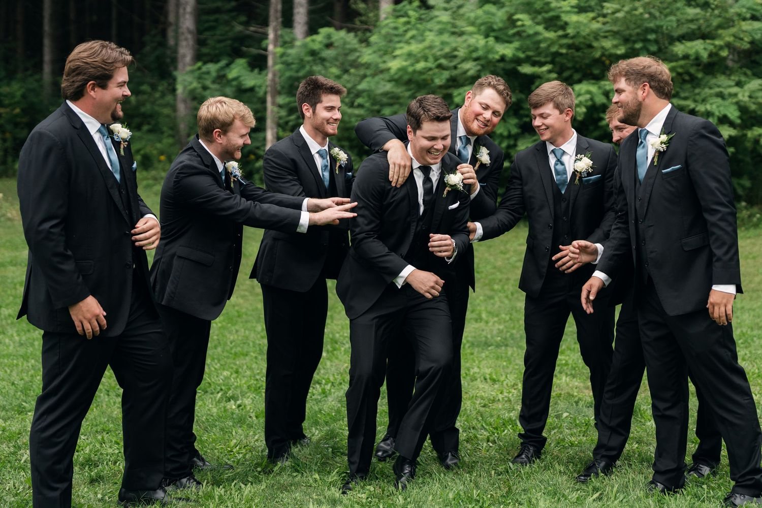 Seven groomsmen in black suits with boutonnieres laughing and joking together outdoors on green grass at a wedding.