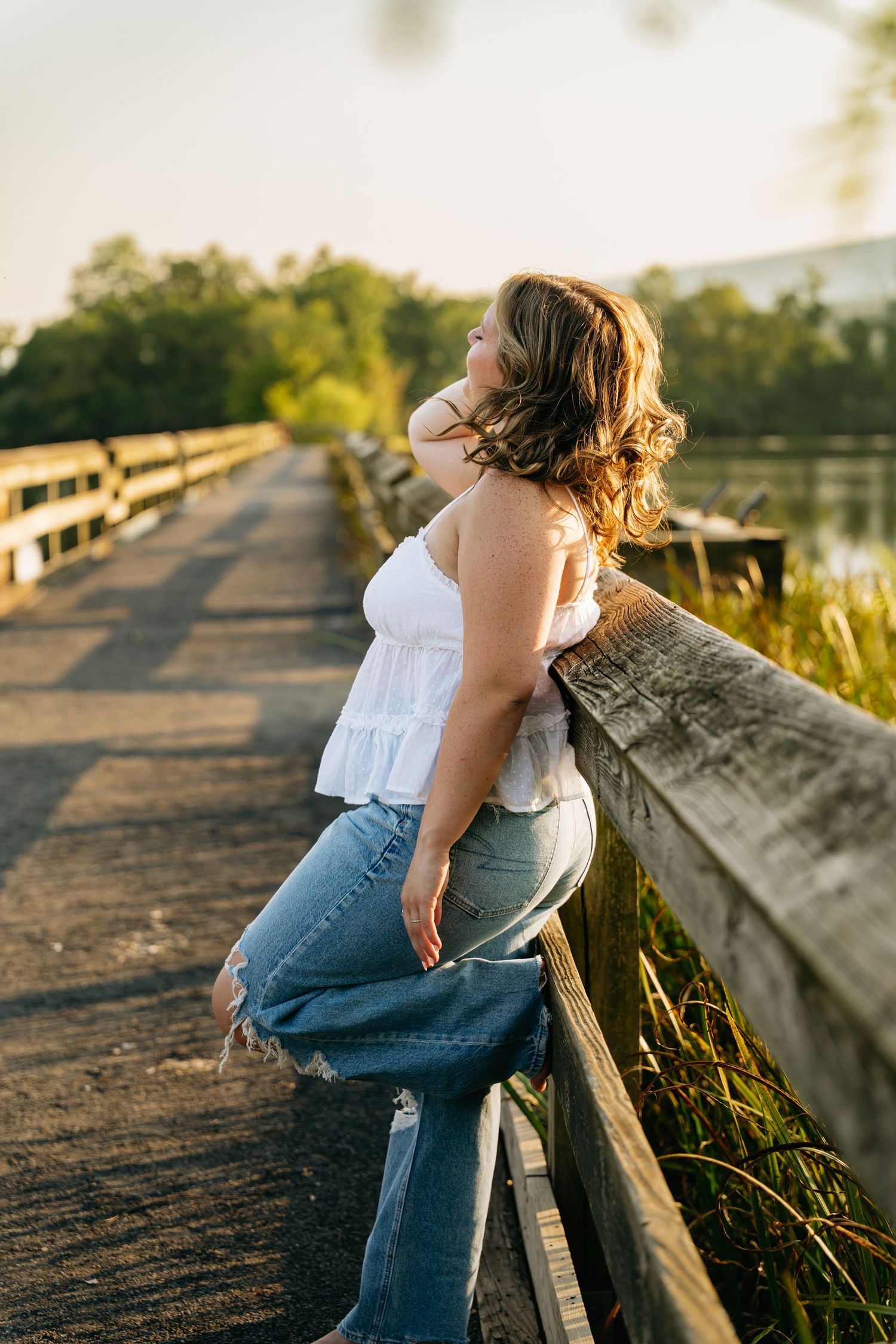 Person in blue jeans and white top leans against wooden railing during golden hour sunset.