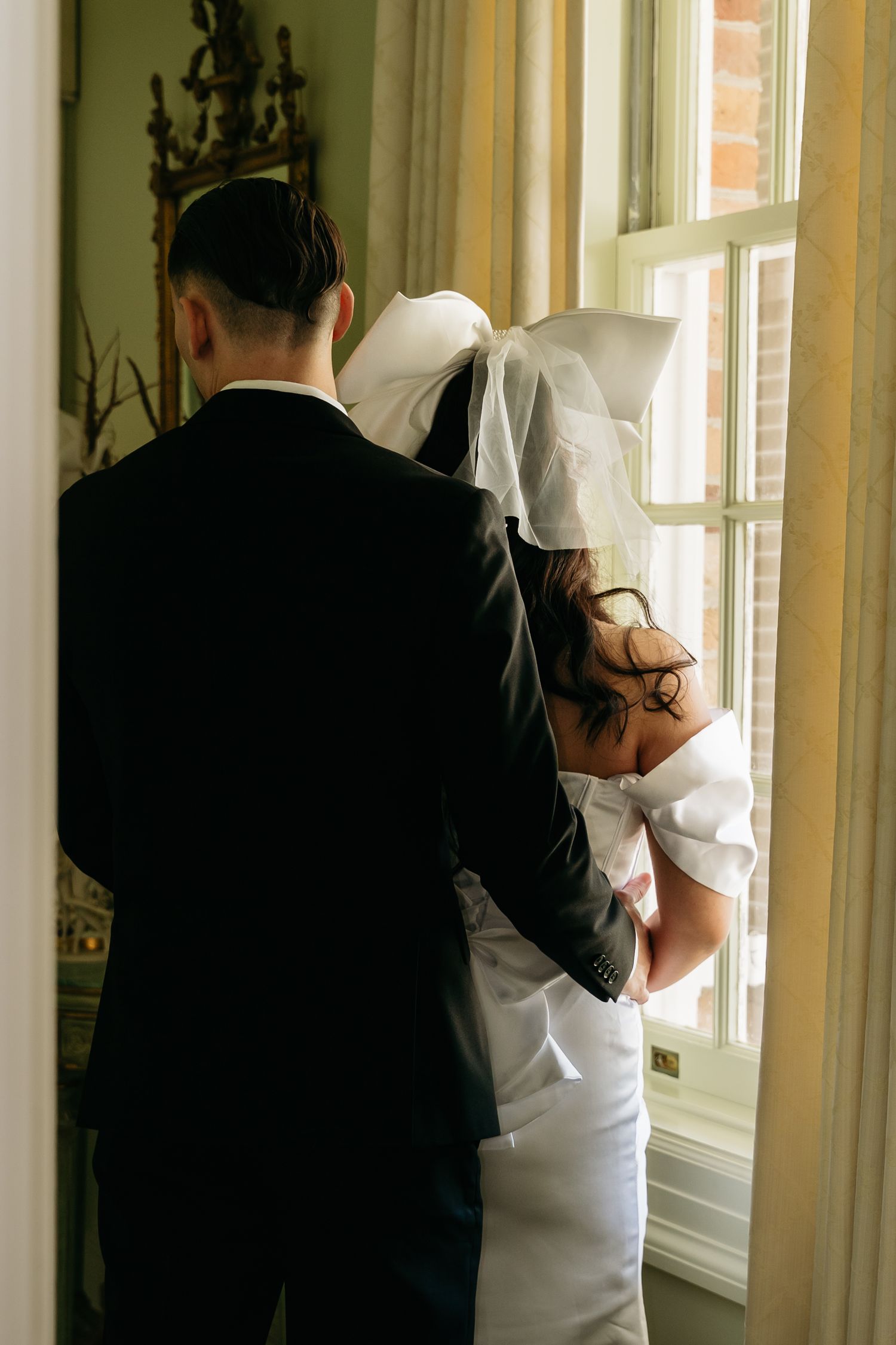 Bride and groom standing together by a window, viewed from behind, wearing wedding attire with veil and black suit.